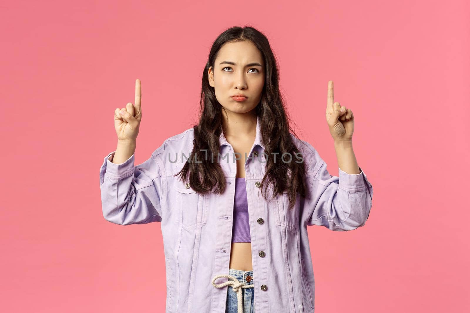 Portrait of gloomy and displeased, young frowning girl in denim jacket, grimacing looking and pointing fingers up disappointed, express regret or jealousy, standing pink background by Benzoix