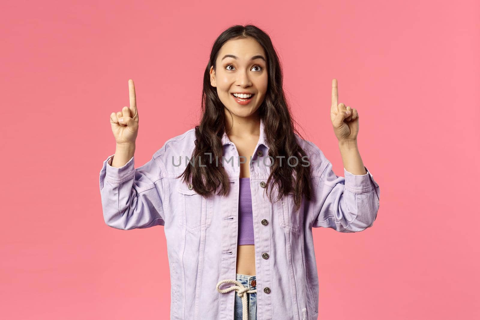 Enthusiastic young pretty girl looking and pointing fingers up with happy smile, see promo, interested in taking part event, party, found great online courses, standing pink background.