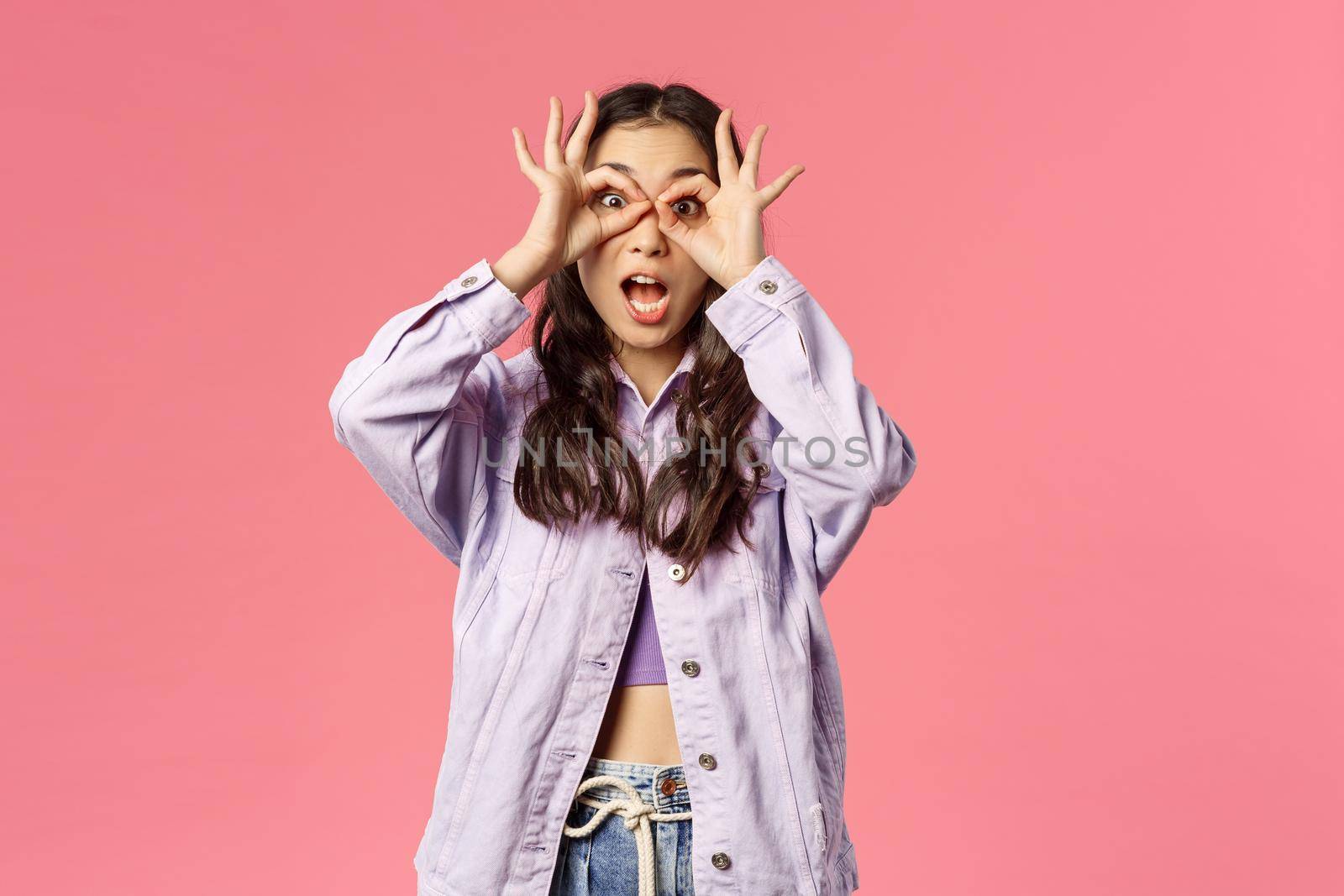 Portrait of funny and silly, playful stylish girl making funny expressions and grimaces, hold hands over eyes like glases or mask, fool around like child, standing pink background by Benzoix