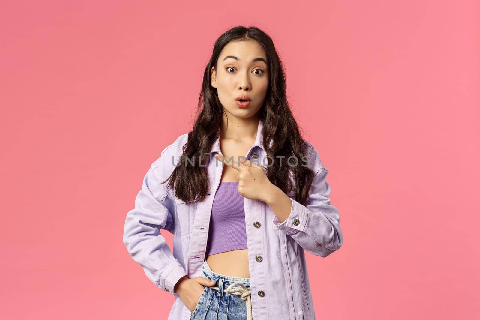 Portrait of surprised and curious young stylish female student pointing finger left and look amazed camera, asking question about interesting thing located there, pink background.