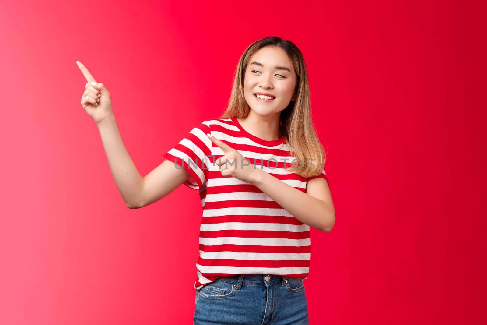 Happy relaxed good-looking outgoing modern urban korean blond girl turn look left pointing fingers upper angle smiling broadly, showing friends thing, stand red background delighted have fun.