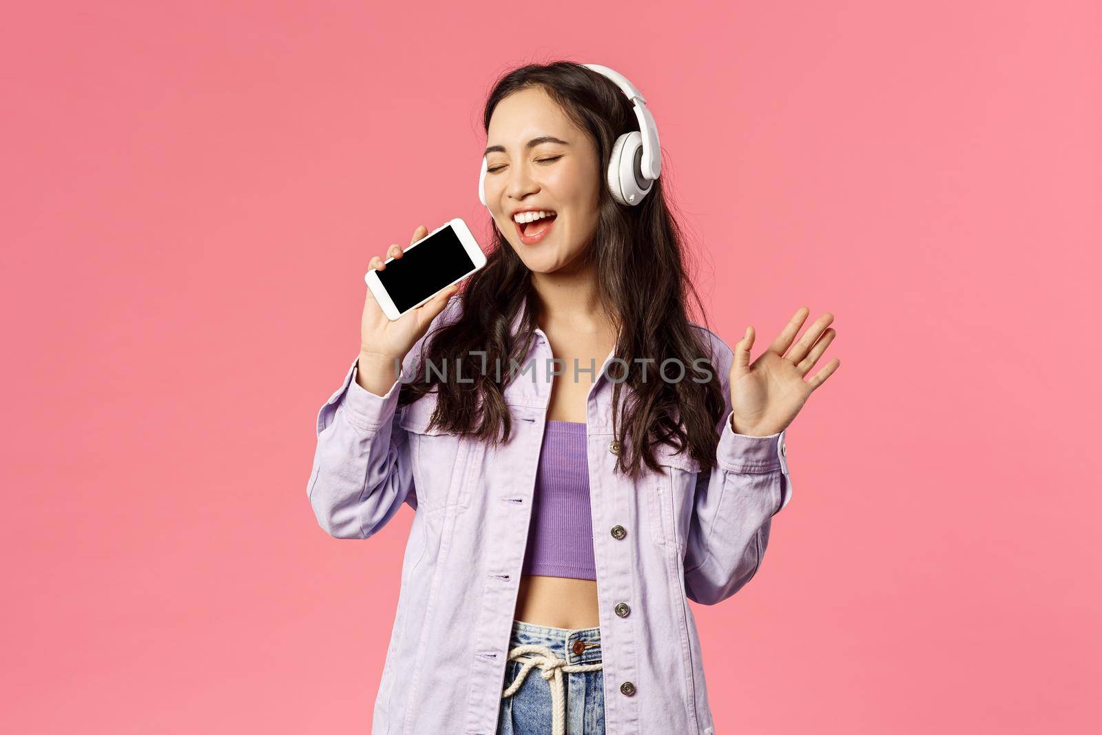 Waist-up portrait of stylish happy woman using headphones to listen awesome new track, download karaoke app and using smartphone like microphone, singing along awesome music by Benzoix