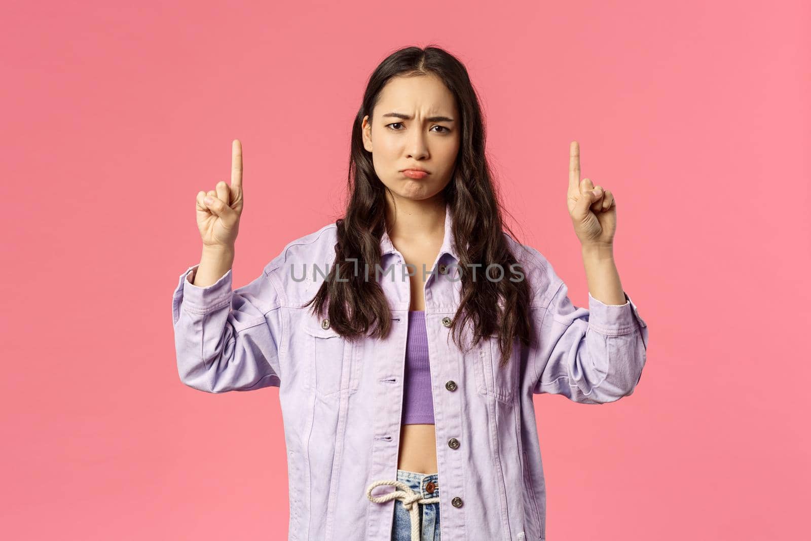 Portrait of uneasy, sulking unhappy asian girl in denim jacket over crop-top, complain unfair situation, pointing fingers up, look camera offended or gloomy, standing pink background by Benzoix