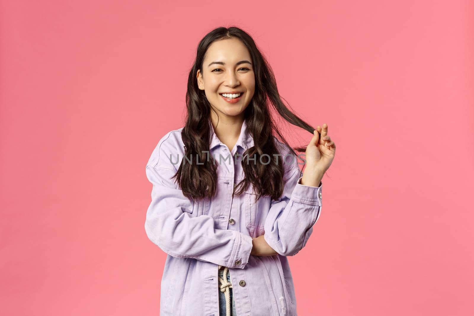 Portrait of cheerful pretty korean woman in denim jacket, roll strand of hair with fingers, giggle and smiling camera, talking to friends, make fashion blog for followers online, over pink background.