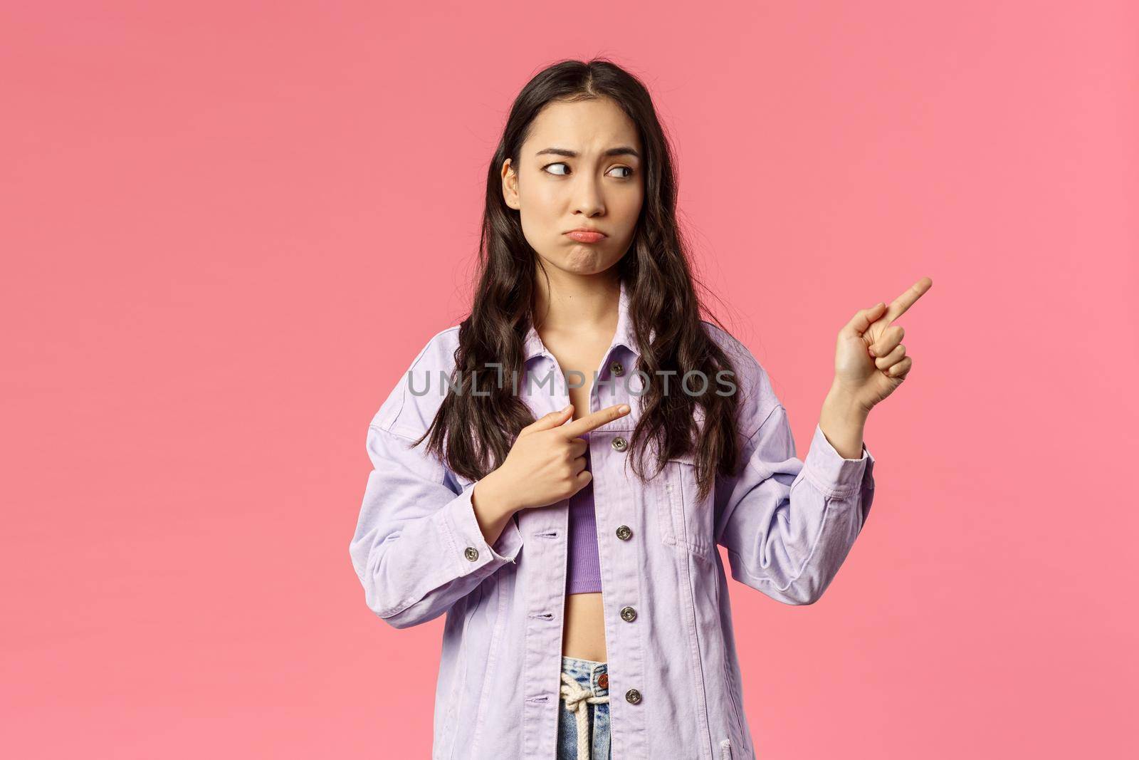 Attractive, gloomy mixed-race girl pointing fingers right, looking at something with regret and sigh, pouting displeased, jealous of missing good opportunity, want be there, pink background by Benzoix