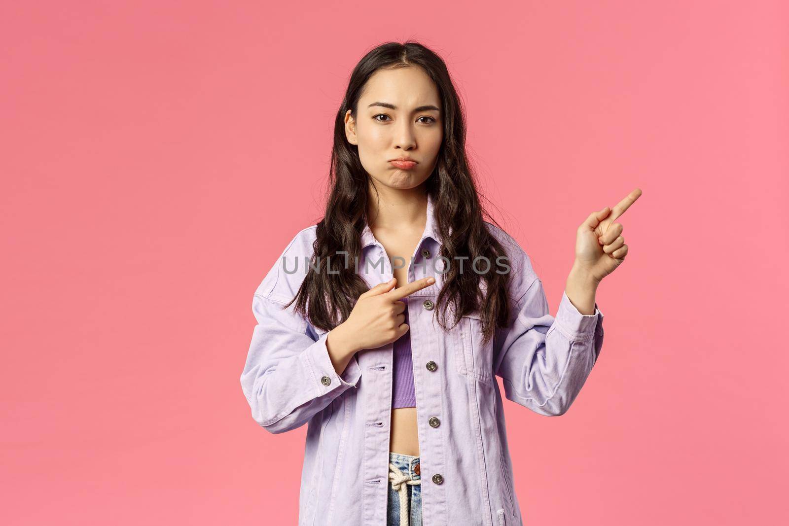 Portrait of envy cute asian girl pouting and looking camera gloomy as pointing fingers right, sulking complaining something unfair happened, standing moody pink background by Benzoix