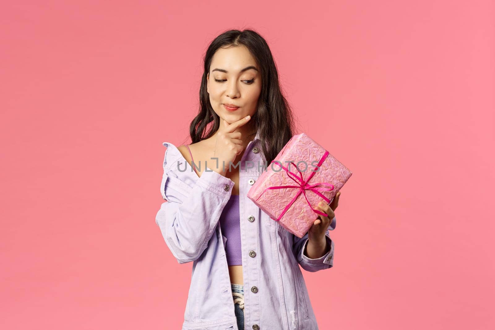 Portrait of curious enthusiastic young asian girl peeking at wrapped box, smirk and touching jaw as thinking about what inside of gift given by friend on birthday party, pink background.