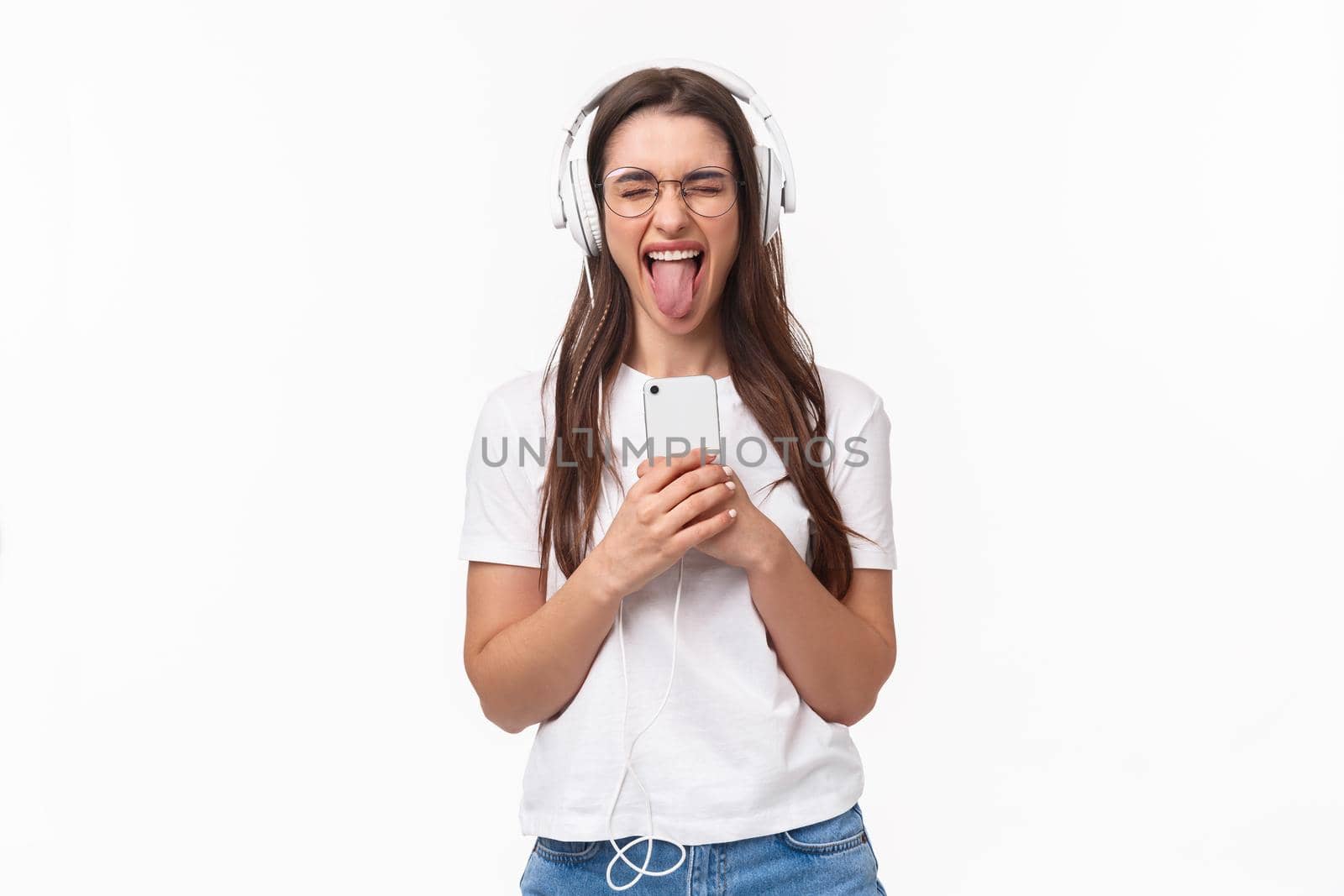 Technology, lifestyle and music concept. Portrait of carefree happy and enthusiastic woman enjoying awesome song, listening in headphones hold smartphone show tongue, white background.