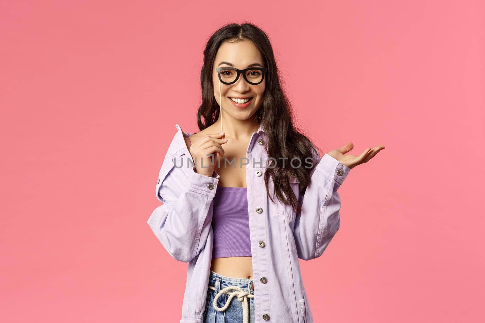 Holidays, lifestyle and people concept. Portrait of good-looking stylish young girl attend party with face mask of glasses on stick, smiling see no proble, standing pink background by Benzoix