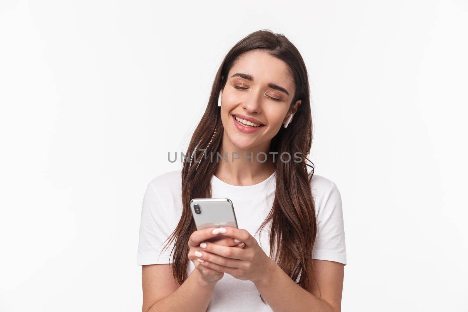 Close-up portrait of tender cute young brunette woman close eyes, shaking hand in rhythm music, smiling from delight, listening song in wireless earphones, holding smartphone.