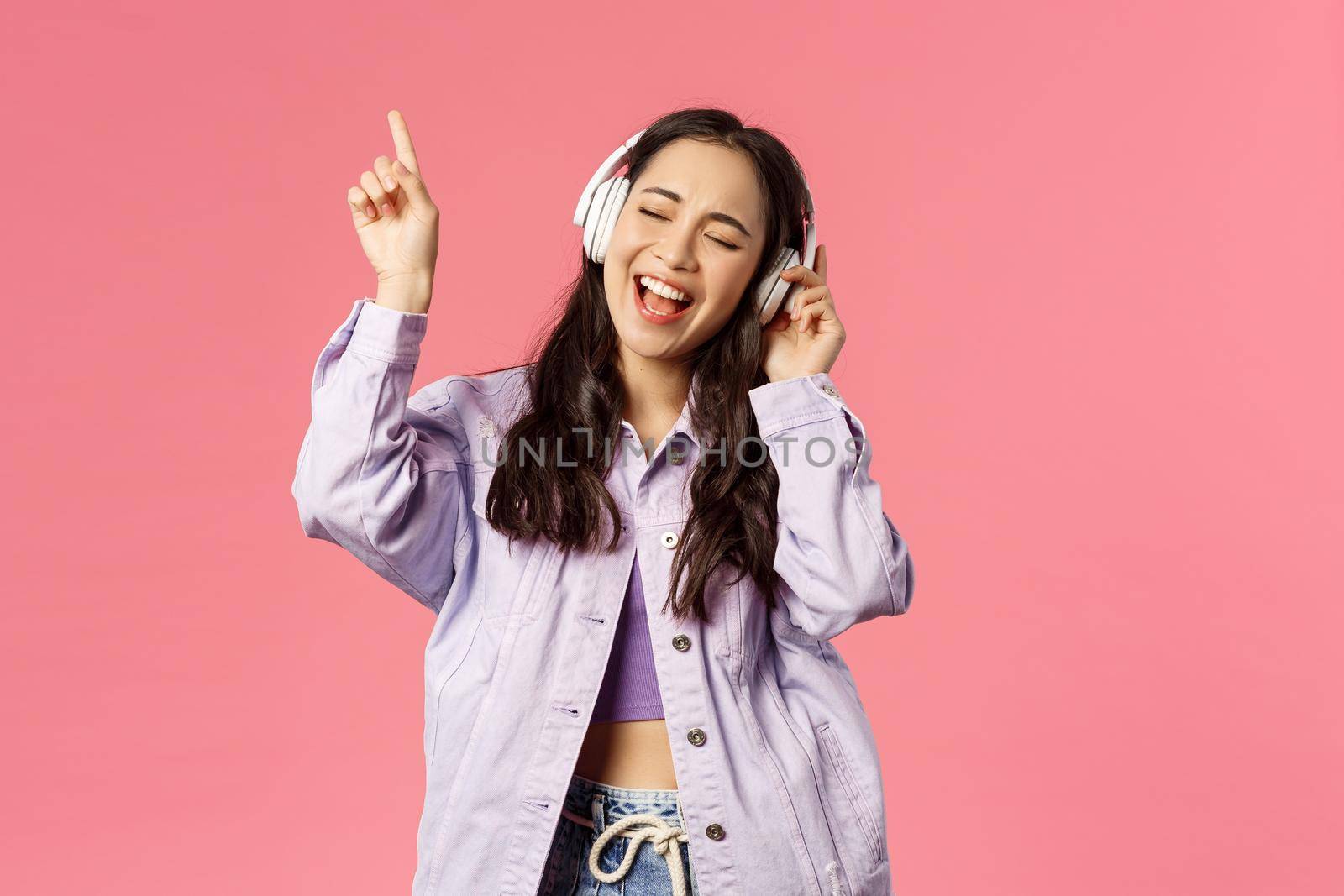 Portrait of attractive carefree asian girl chilling and vibing, listening music in headphones, raising finger up while trying sing along and reach highest note in song, pink background by Benzoix