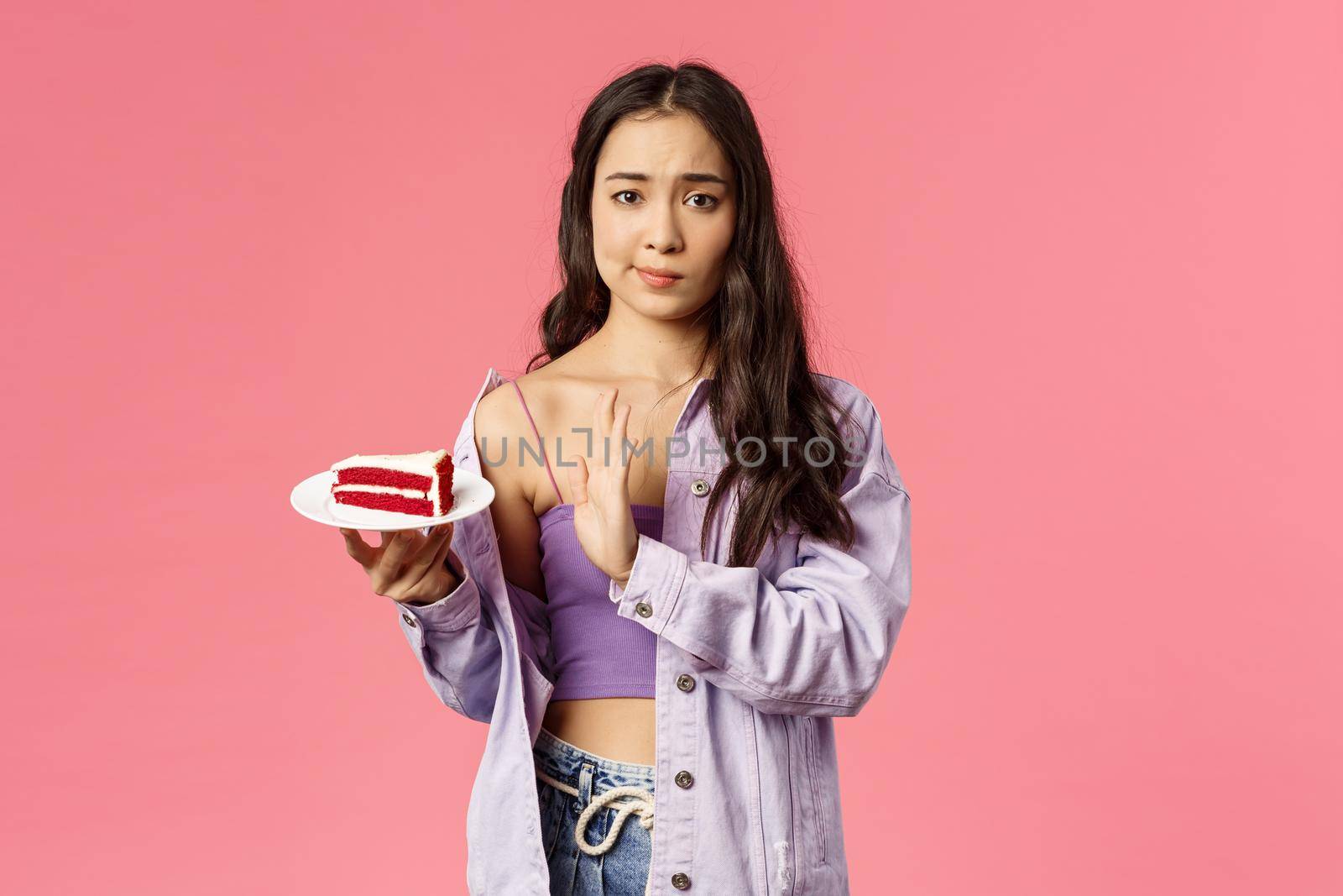 Portrait of skeptical young asian stylish slim girl refusing eat cake, smirk show top sign to dessert lying on plate, rejecting try it, disappointed with bad taste, stand pink background by Benzoix
