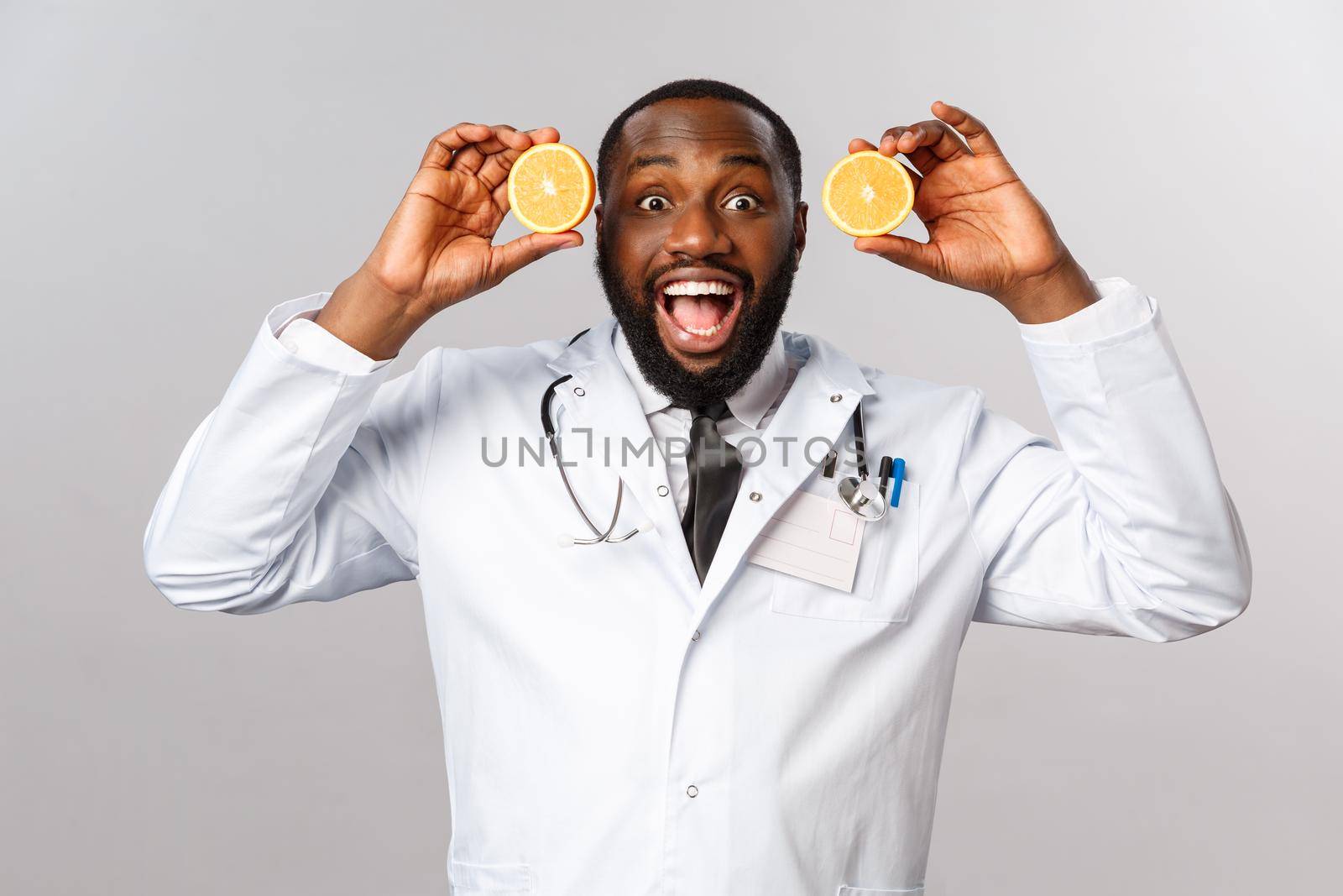 Healthcare, healthy diet and disease concept. Excited handsome african-american doctor in white coat, showing pieces of oranges and smiling amused, advice eat more food consist vitamin c, fruits by Benzoix