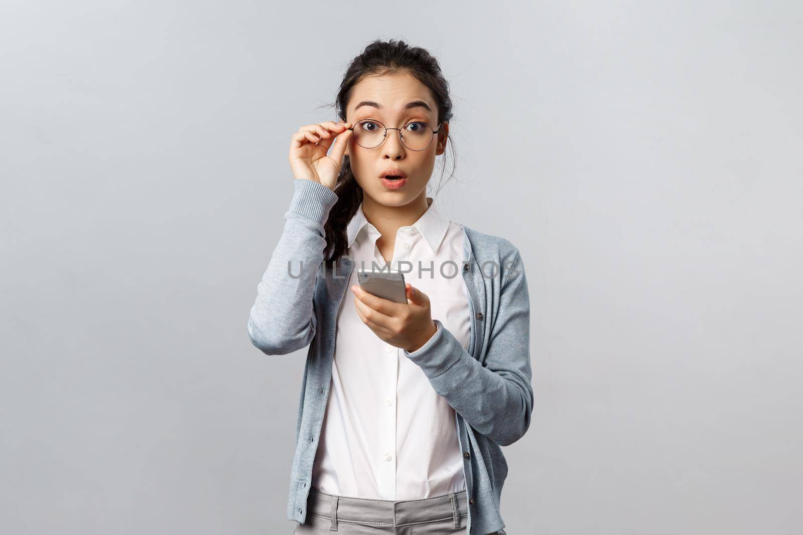 Office lifestyle, business and people concept. Very good. Impressed young asian female fixing glasses on nose, look surprised and amazed camera as using new app on mobile phone or receive text by Benzoix