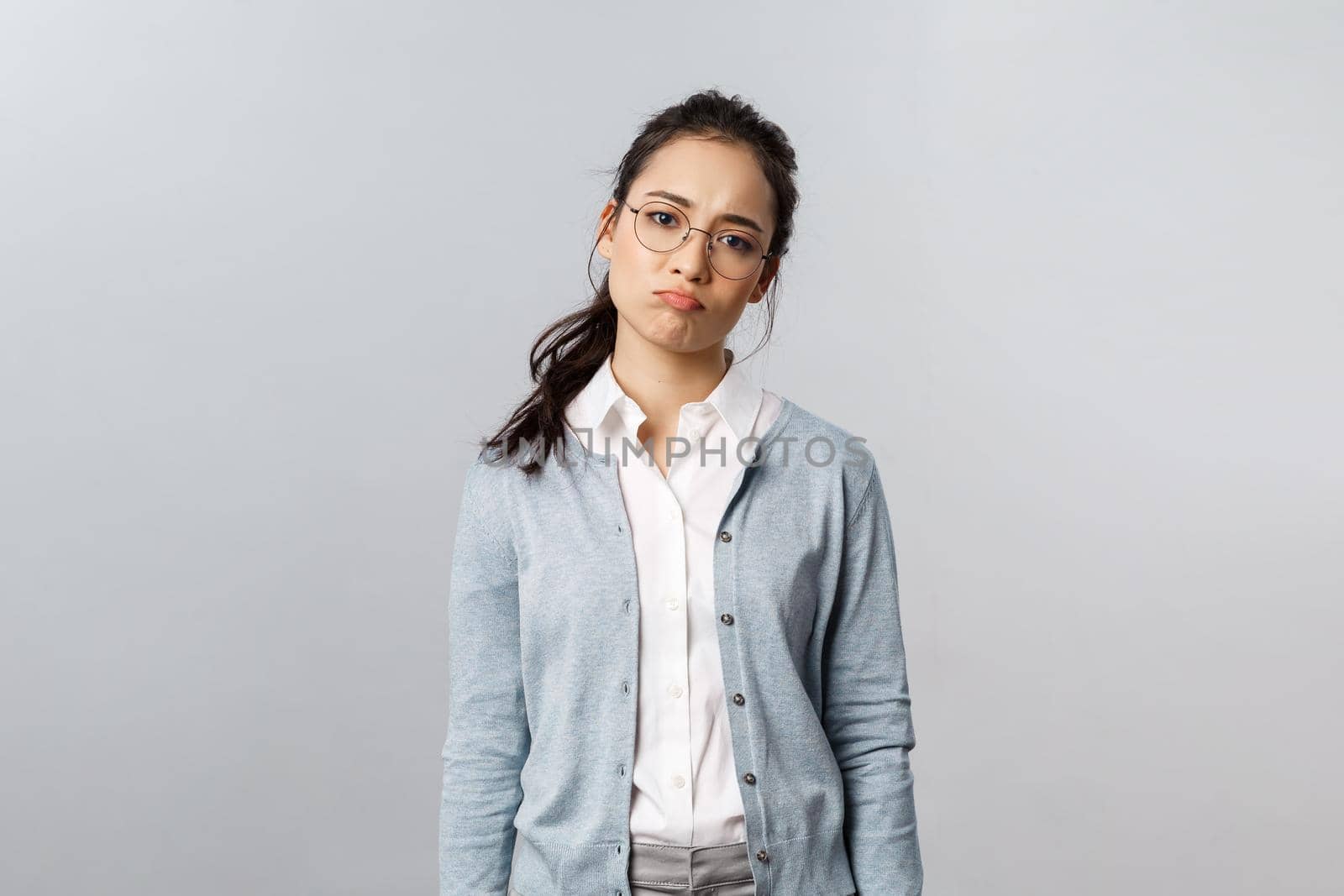 Portrait of sighing displeased and gloomy young asian girl in glasses, pouting and looking disappointed camera, express regret or feel uneasy over failure at work, grey background.