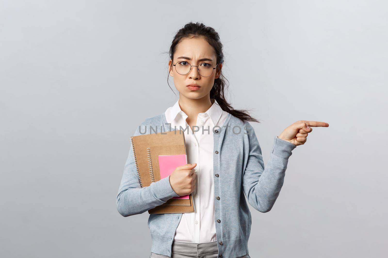 Education, teachers, university and schools concept. Go there, leave now. Serious-looking disappointed frowning female tutor look displeased, pointing finger right, standing grey background by Benzoix