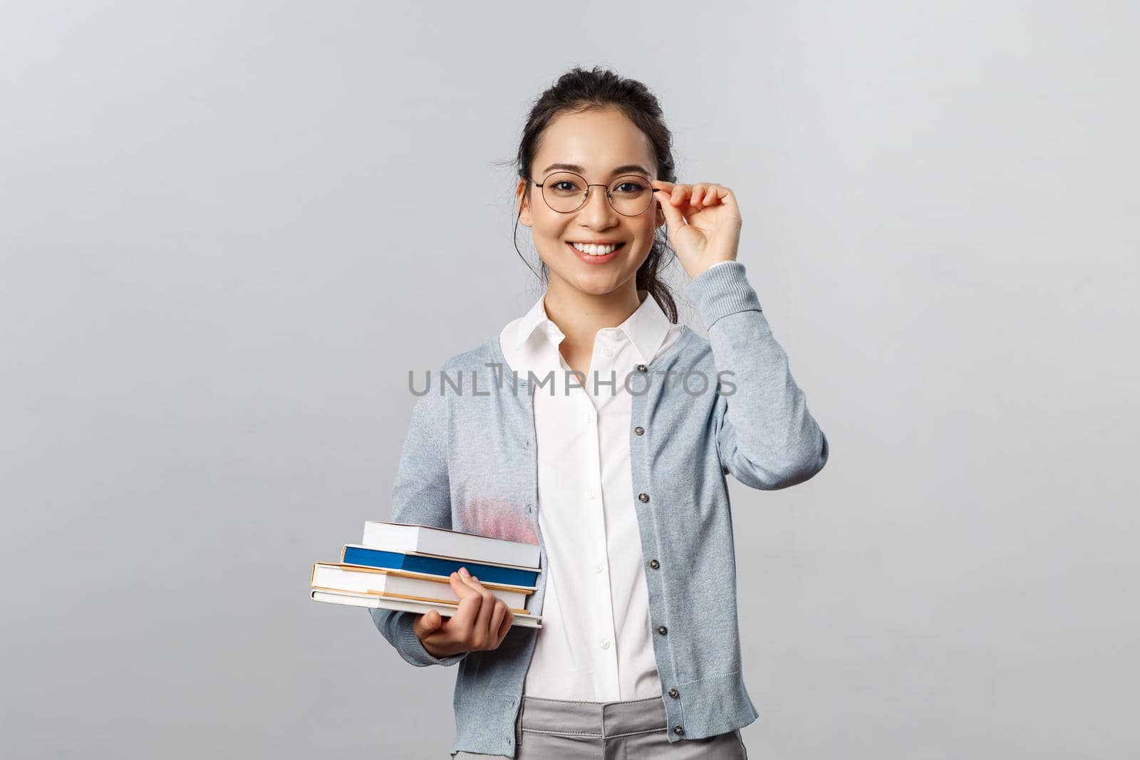 Education, teachers, university and schools concept. Young female student, asian girl studying for exams, fixing glasses on eyes and smiling, carry books to library, stand grey background by Benzoix