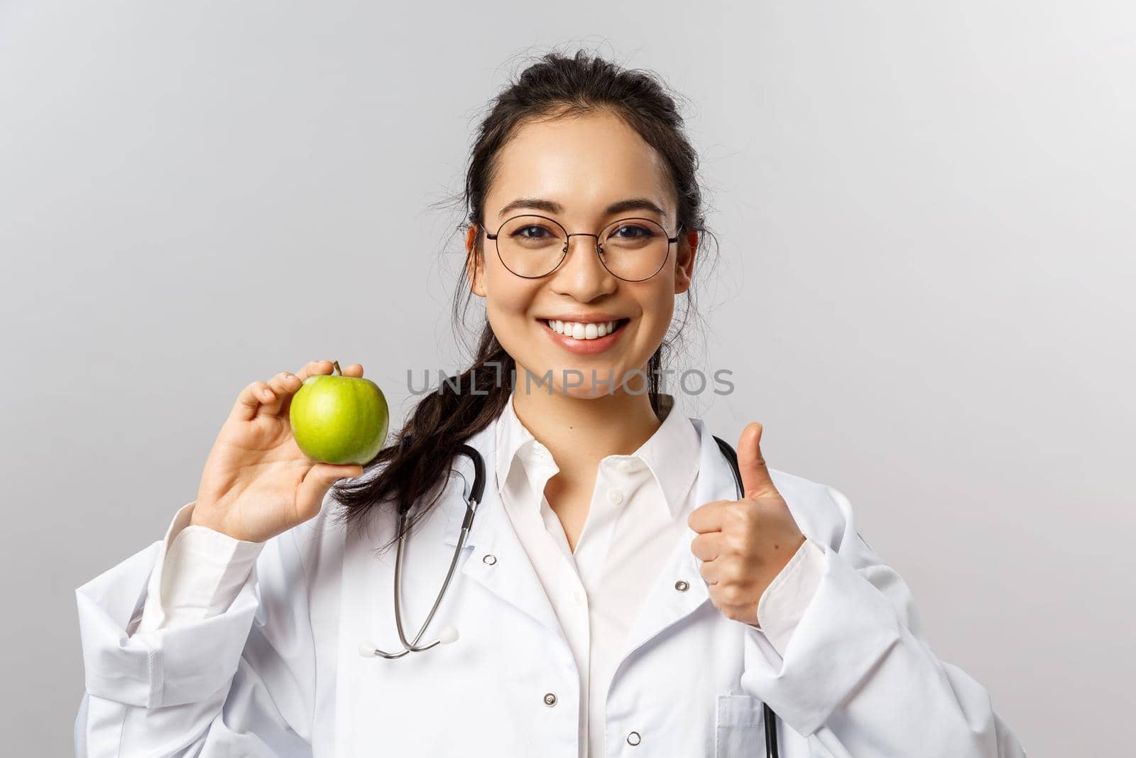 Portrait of young cheerful asian female doctor, therapist or diatologist talking abut healthy diet, showing green apple and thumb-up, recommend eat fruits, smiling encourage look after health by Benzoix