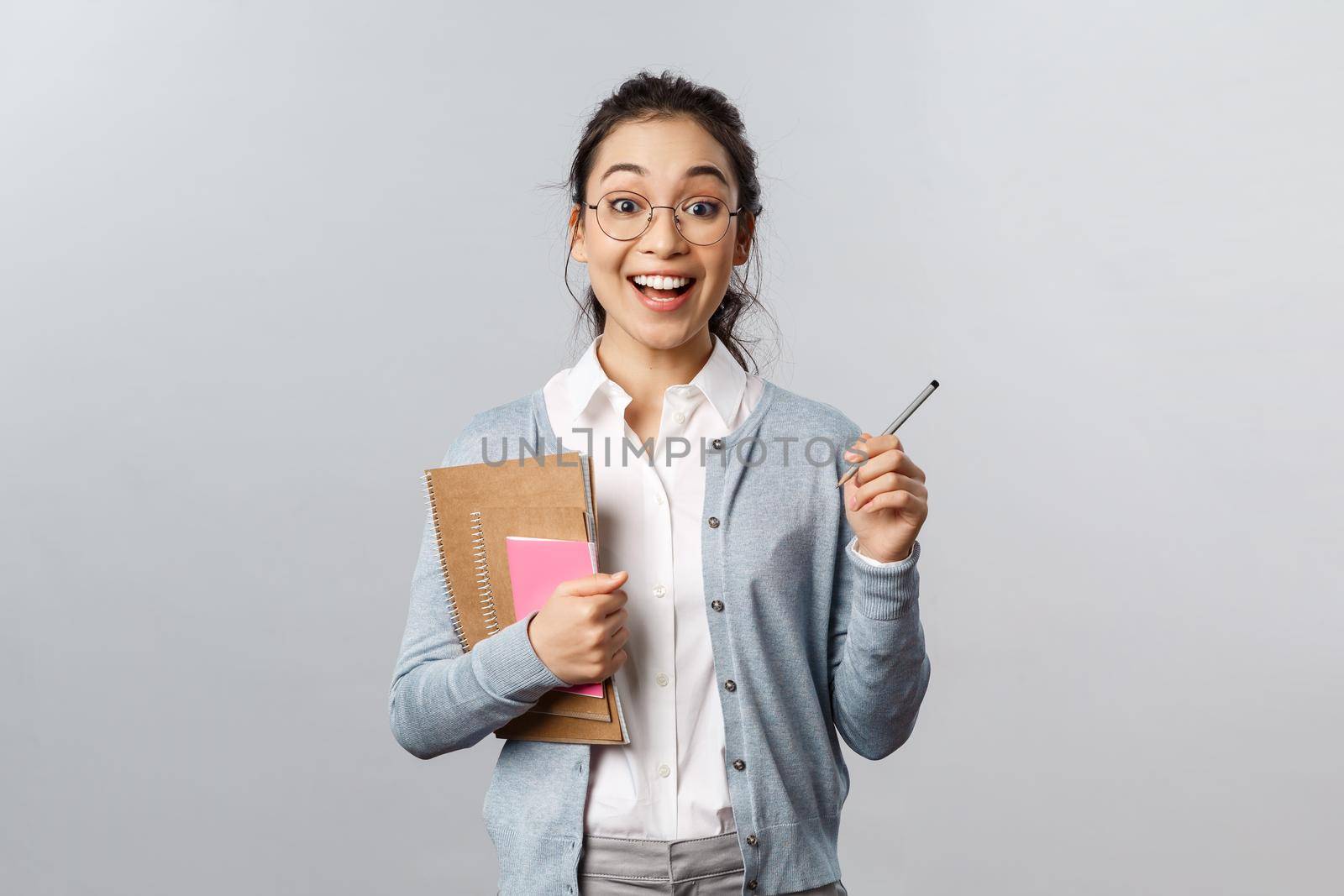 Education, teachers, university and schools concept. Enthusiastic young asian female in glasses, holding pencil and notebooks, write good mark at student essay, pleased with great homework by Benzoix