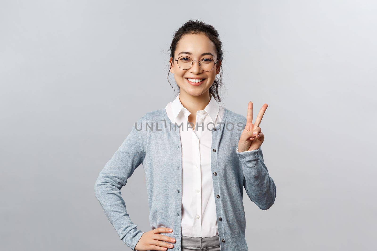 People, emotions and lifestyle concept. Upbeat optimistic asian woman in glasses, school teacher or tutor show peace sign and smiling friendly, express positive cheerful emotion, grey background.