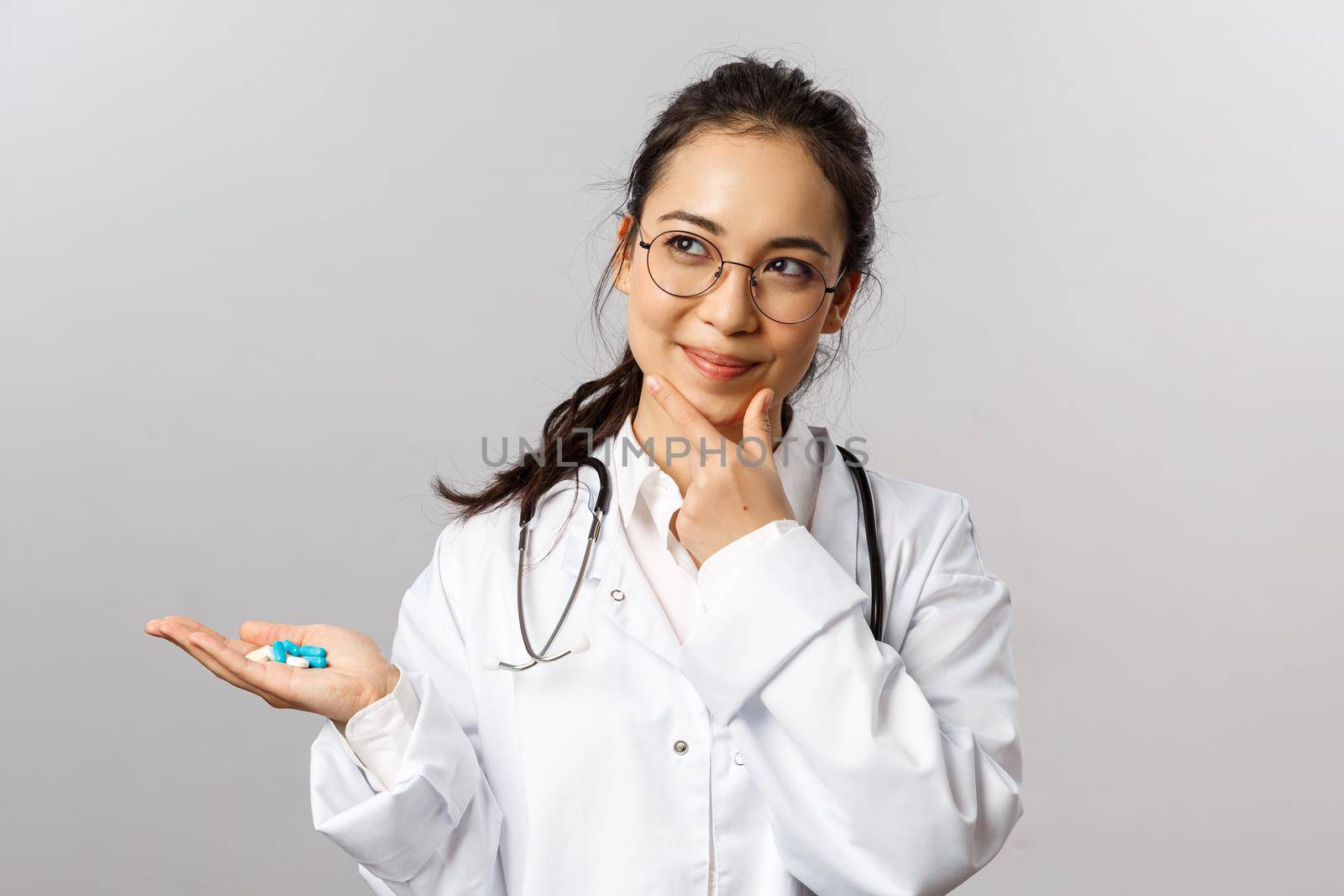 Healthcare, medicine and pharmacy concept. Portrait of thoughtful, pleased female asian doctor, smirk and look up, thinking satisfied, holding pills, invent new drugs to cure sick people.