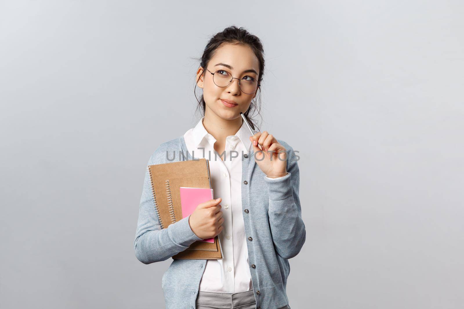 Education, teachers, university and schools concept. Thoughtful asian female student, solving puzzle, look up with intrigued, thinking face, bite pencil while pondering, hold notebooks by Benzoix