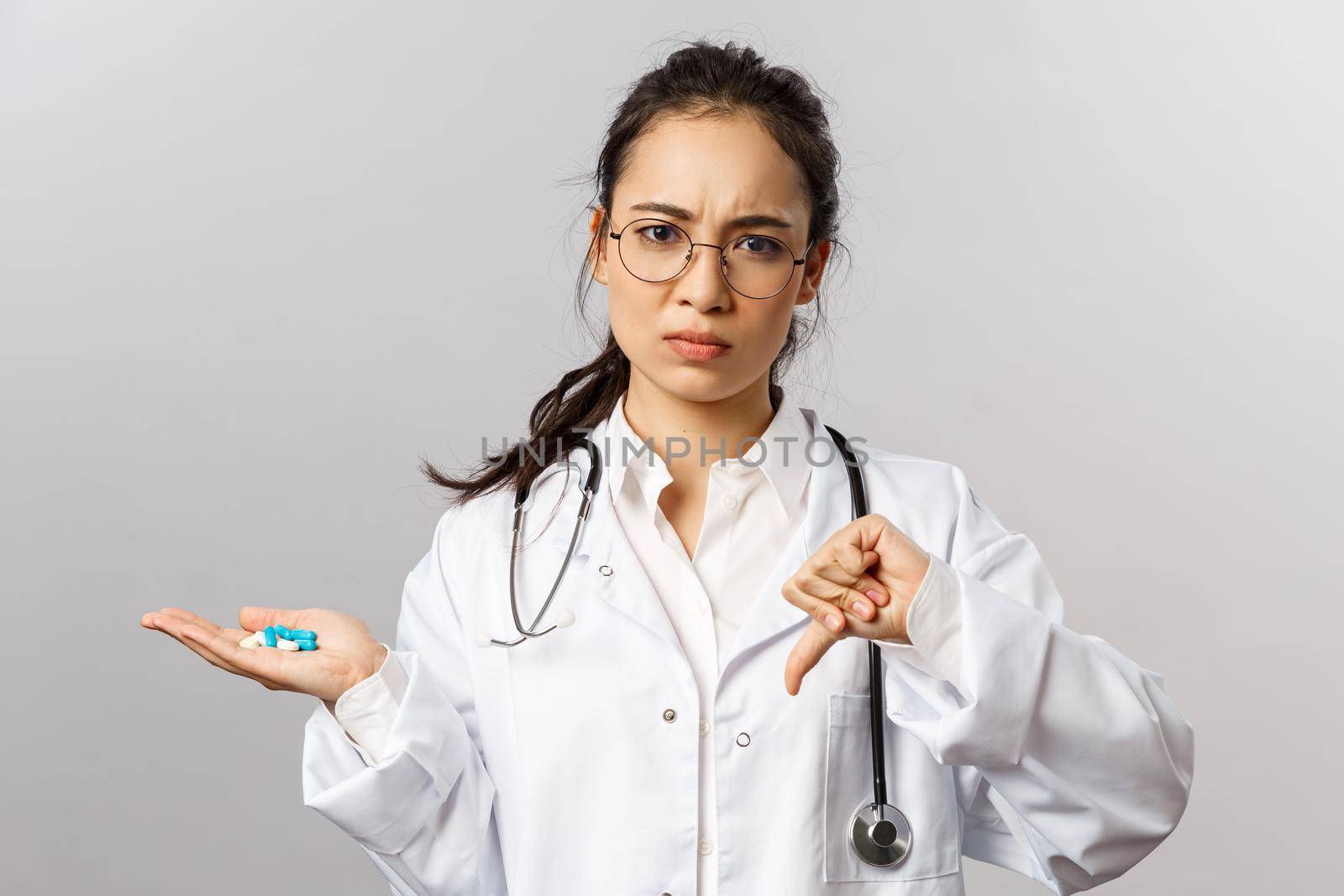 Healthcare, medicine and pharmacy concept. Portrait of worried asian female doctor, frowning disturbed, thumbs-down and hold bad pills, advice not to self-medicate from virus, contact your therapist by Benzoix