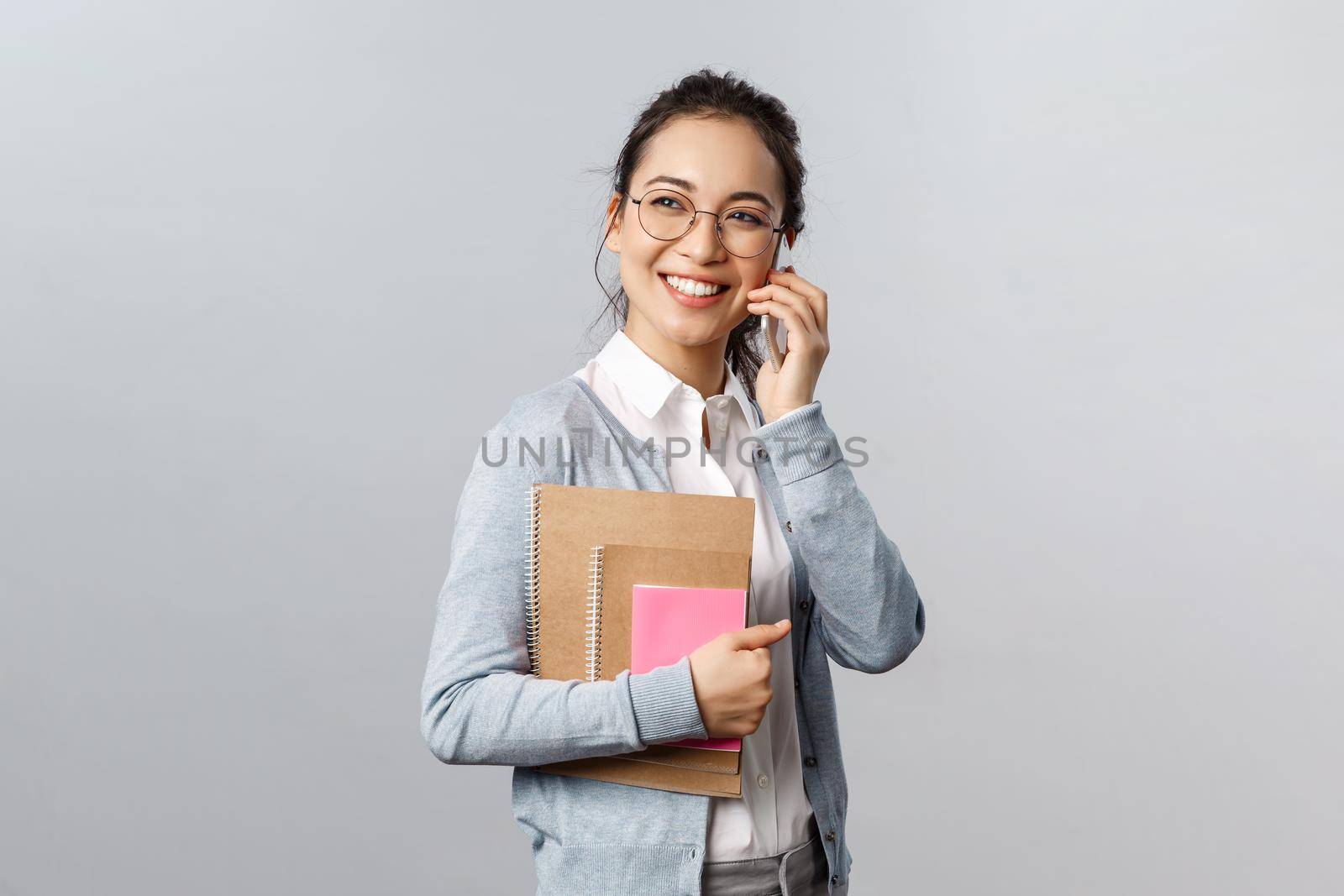 Education, teachers, university and schools concept. Cheerful smiling asian woman, tutor answer phone call, talking with friend laughing and looking away pleased, carry notebooks.
