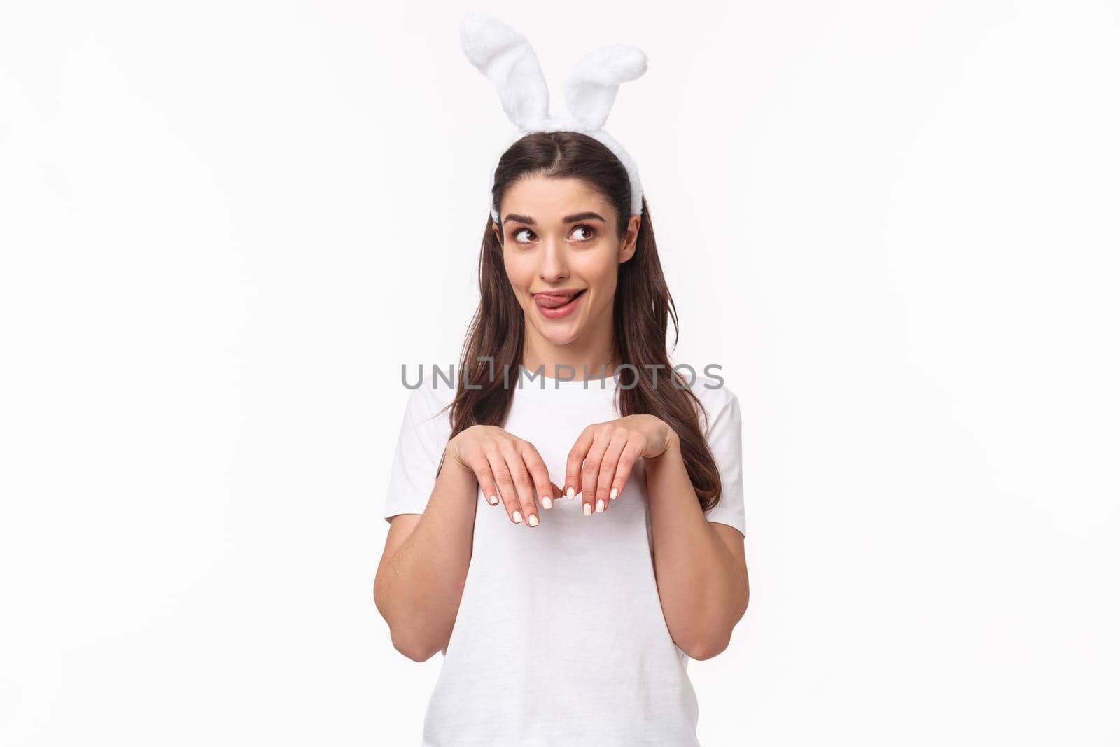 Funny and silly, playful girl in rabbit ears, t-shirt, looking away and licking lips, daydreaming about something delicious on Easter day, imitating bunny with hands pulled close to chest like paws.