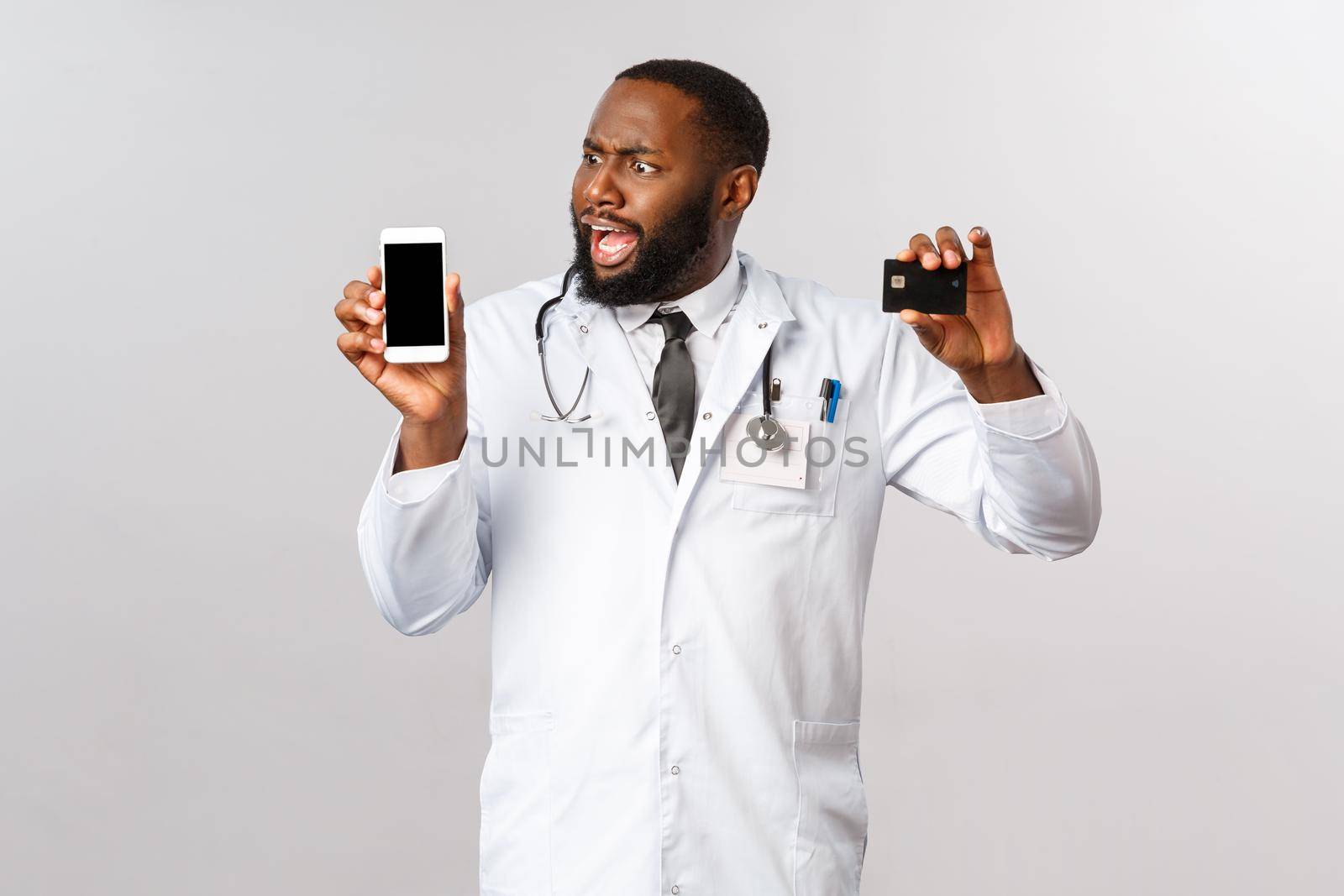 Covid19, pandemic and online medicine concept. Complaining angry african-american man looking at mobile phone display, hold credit card, outraged with huge bills and expensive app subscribtion.