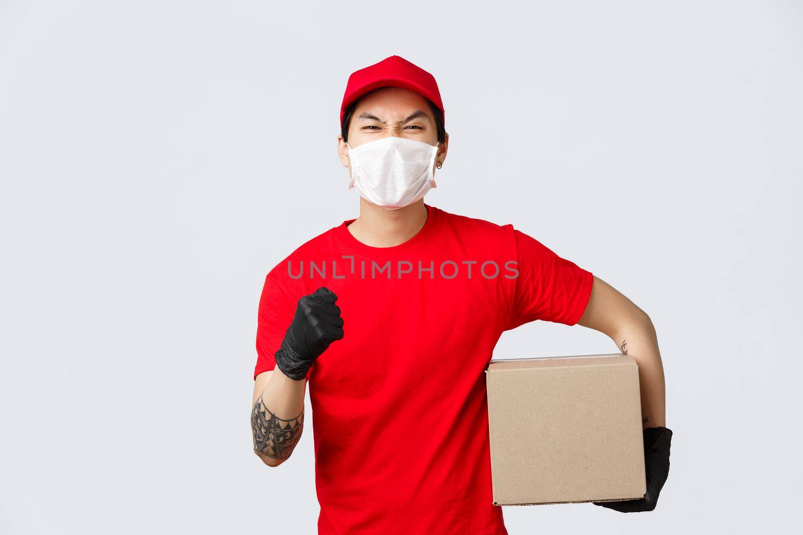 Deliver your goods no matter what. Confident asian delivery guy in red uniform cap and t-shirt, raise hand encourage place your order, holding box pachage, wear medical mask and gloves.