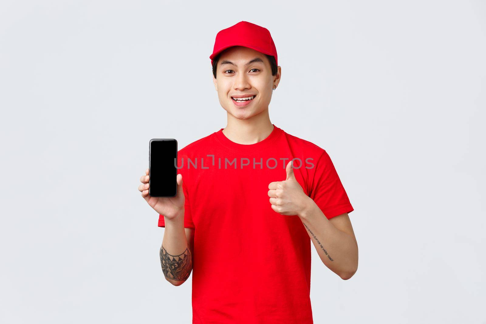 Delivery application, online shopping and shipping concept. Handsome asian courier in red t-shirt and cap, show thumb-up in approval and mobile phone screen app, provide fast and quality service.