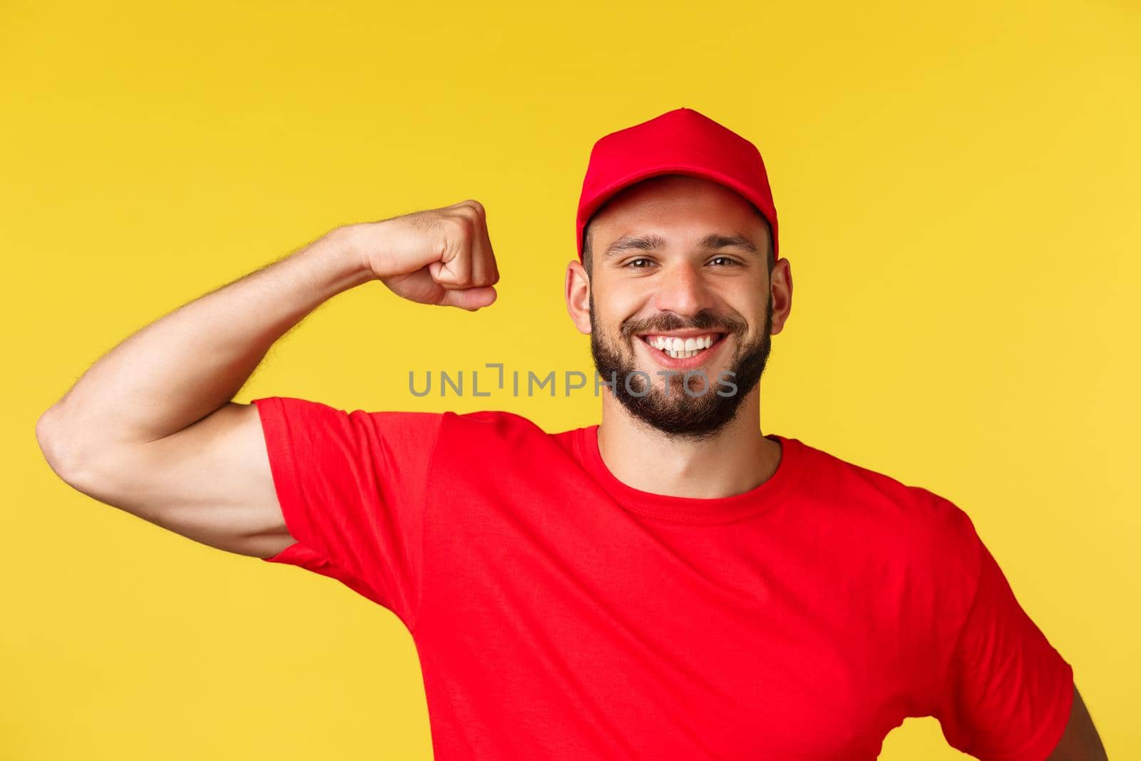 Close-up of confident and cheerful delivery guy showing how storng he is, can carry huge heavy packages, flex piceps and smiling pleased. Courier wearing red uniform over yellow background.