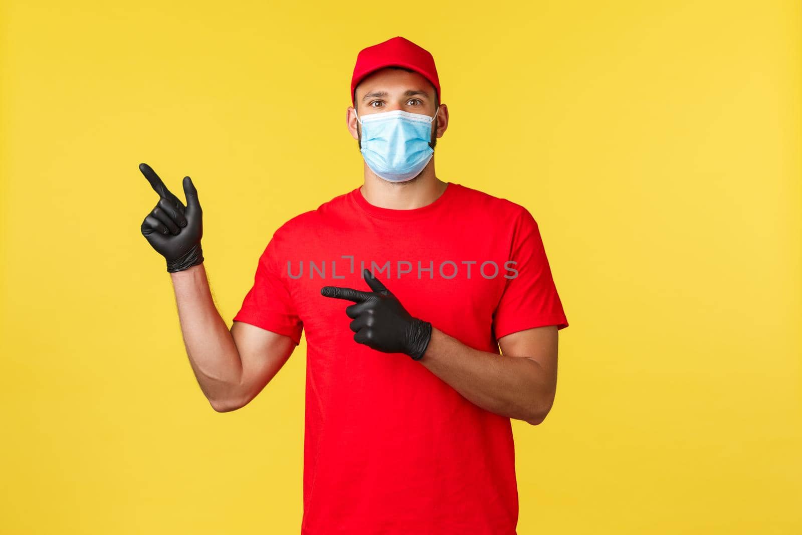Express delivery during pandemic, covid-19, safe shipping, online shopping concept. Enthusiastic courier in red uniform, medical mask and gloves, pointing fingers left, inform about promo.