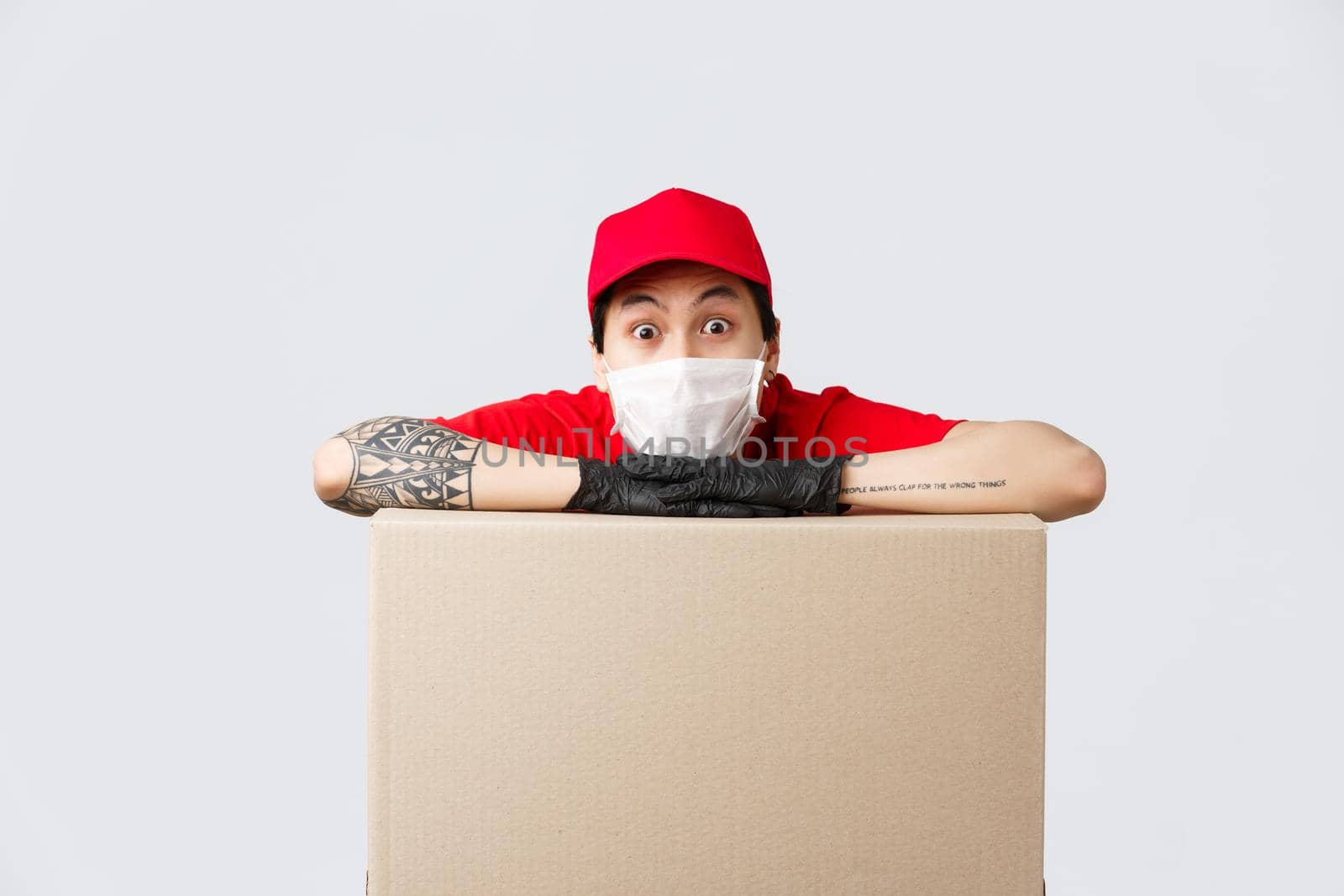 Excited asian delivery guy in medical mask and protective gloves, leaning on box parcel and staring camera astonished. Courier working during coronavirus pandemic, safely deliver orders.