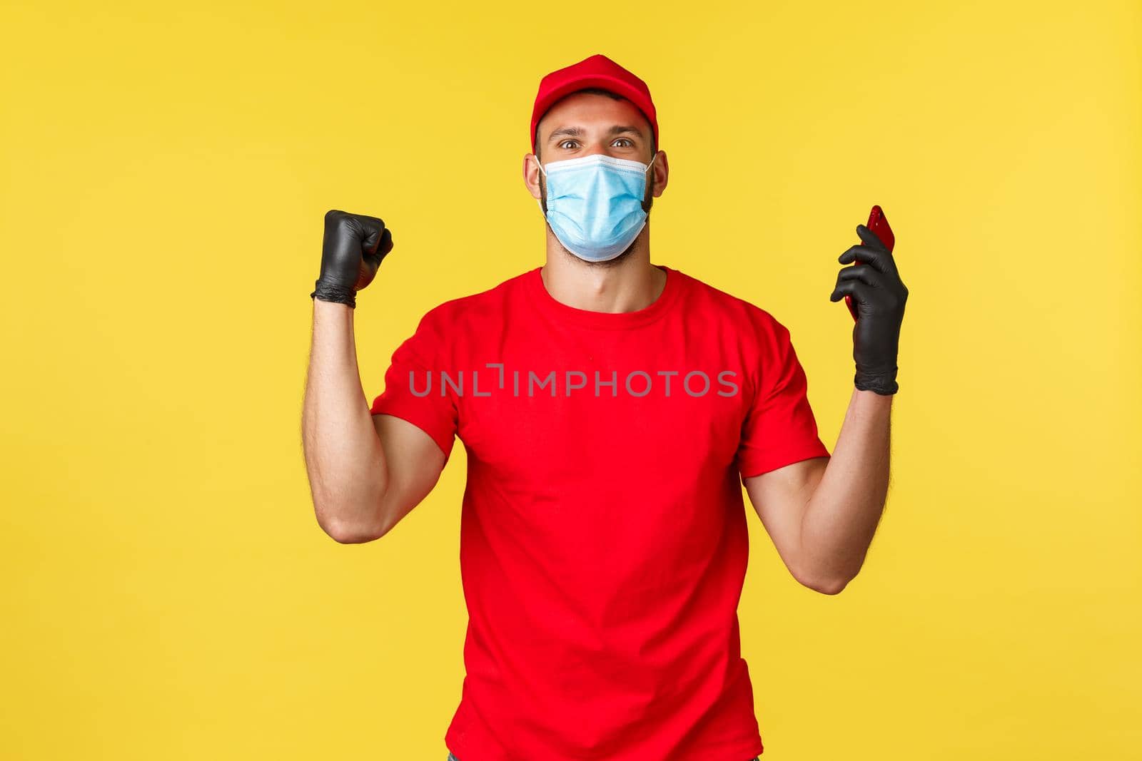 Express delivery pandemic, covid-19, safe shipping, online shopping concept. Satisfied excited courier in red uniform, medical mask and gloves, chanting, celebrating good news, hold mobile phone.