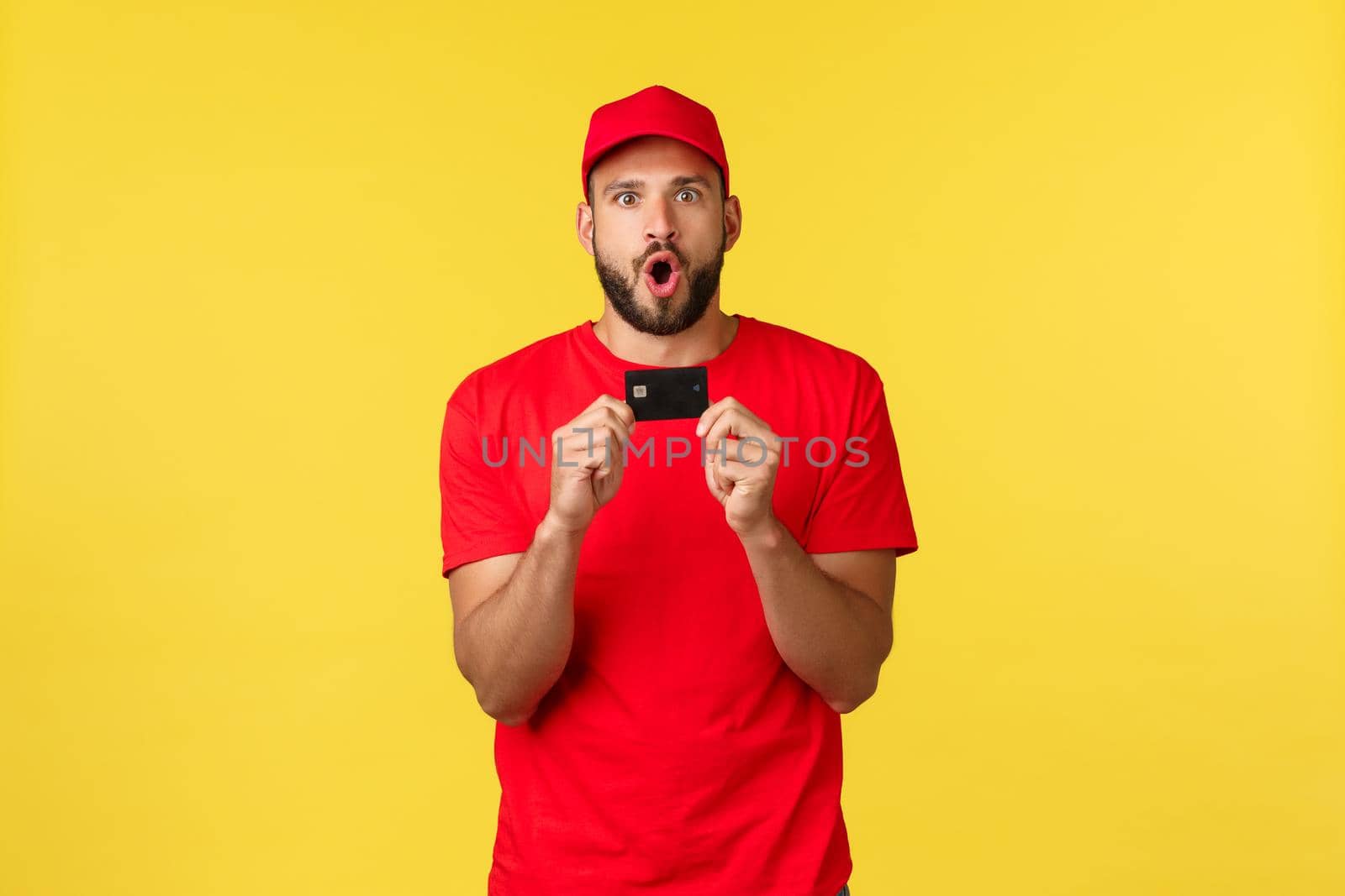 Contactless payment, online delivery, internet shopping concept. Excited and amazed young male employee in company uniform, red t-shirt and cap telling amazing news, show credit card.