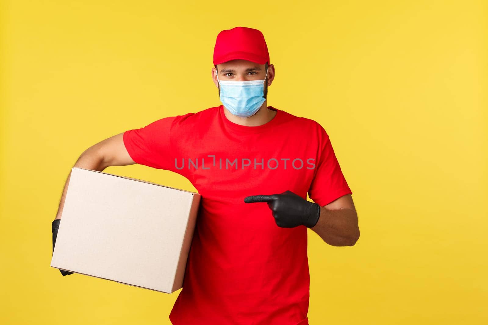 Express delivery during pandemic, covid-19, safe shipping, online shopping concept. Handsome courier in medical mask, red uniform and gloves asking where put package, pointing at box.
