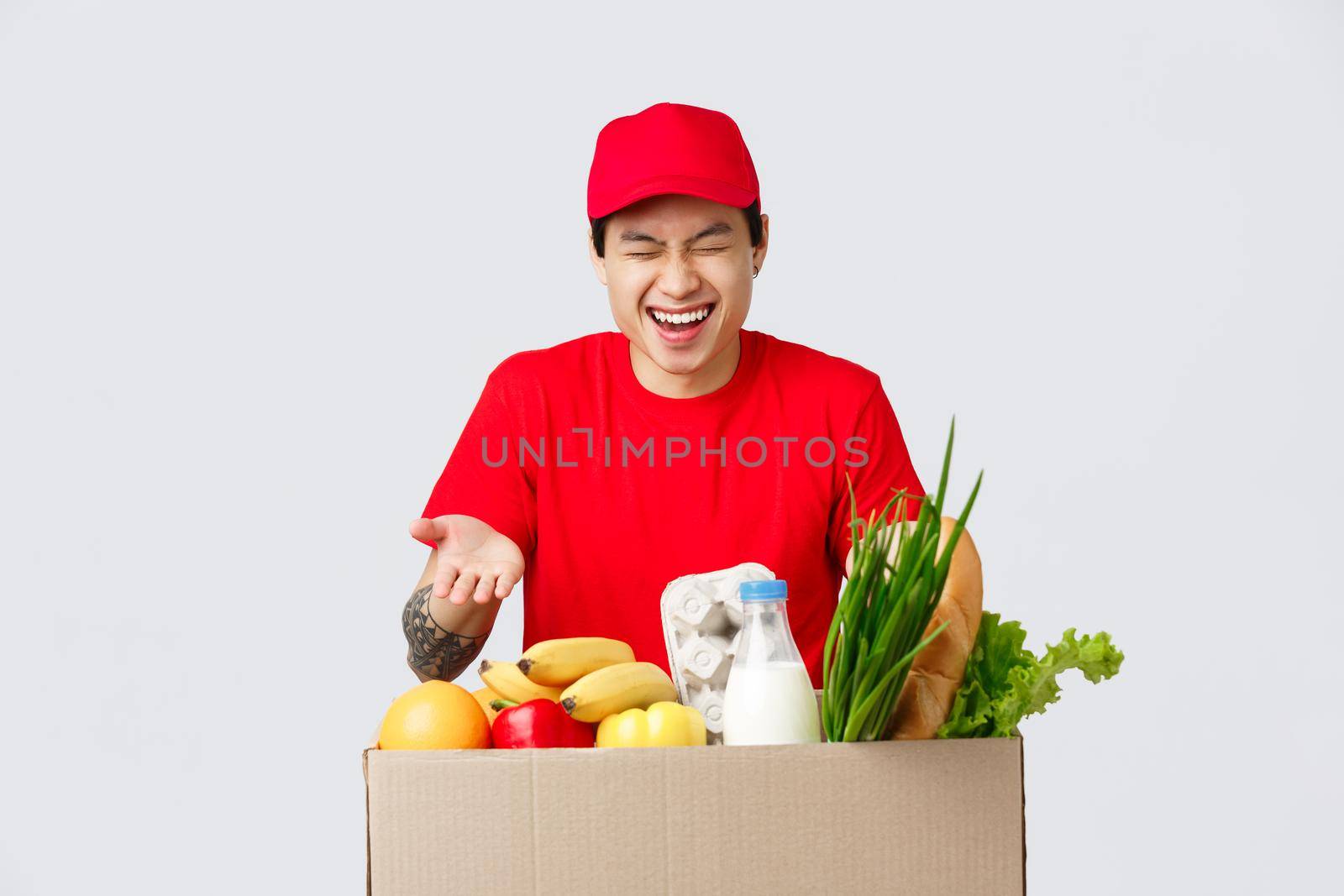 Online shopping, food delivery and internet stores concept. Handsome asian male courier in red uniform, laughing as joking with employees, deliver grocery packages to customer placed order.