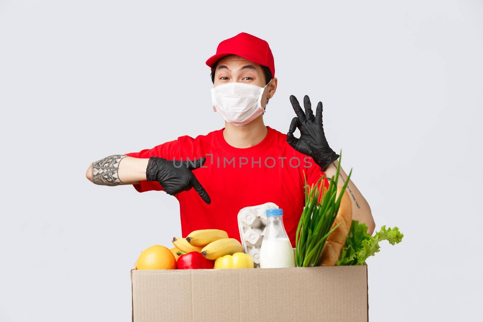 Online shopping, food delivery and coronavirus pandemic concept. Excellent courier service provided by asian delivery man, show okay sign guarantee quality, pointing package with groceries, wear mask.