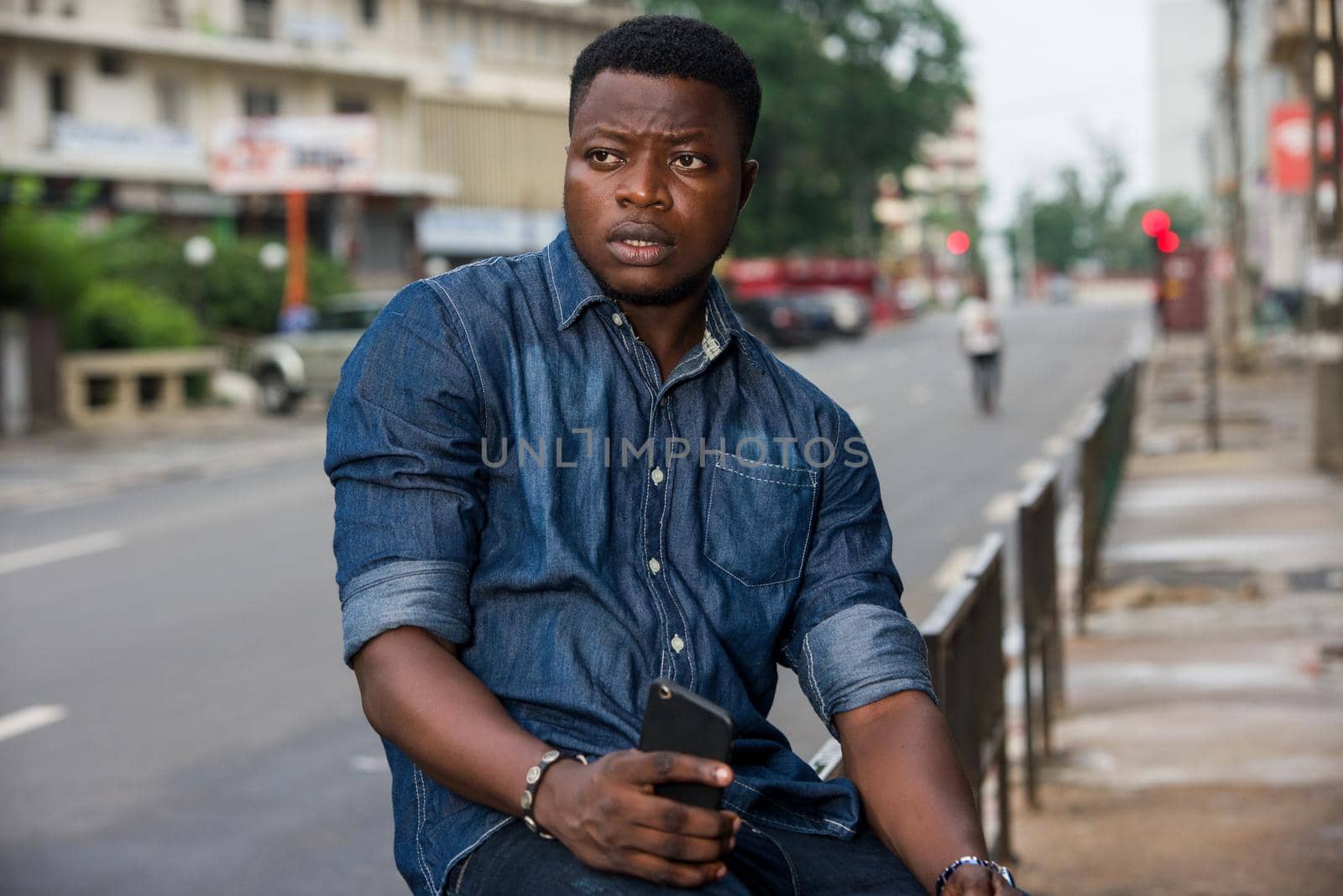 Confident businessman or manager sitting on an iron bar, holding a cell phone and looking aside in the city