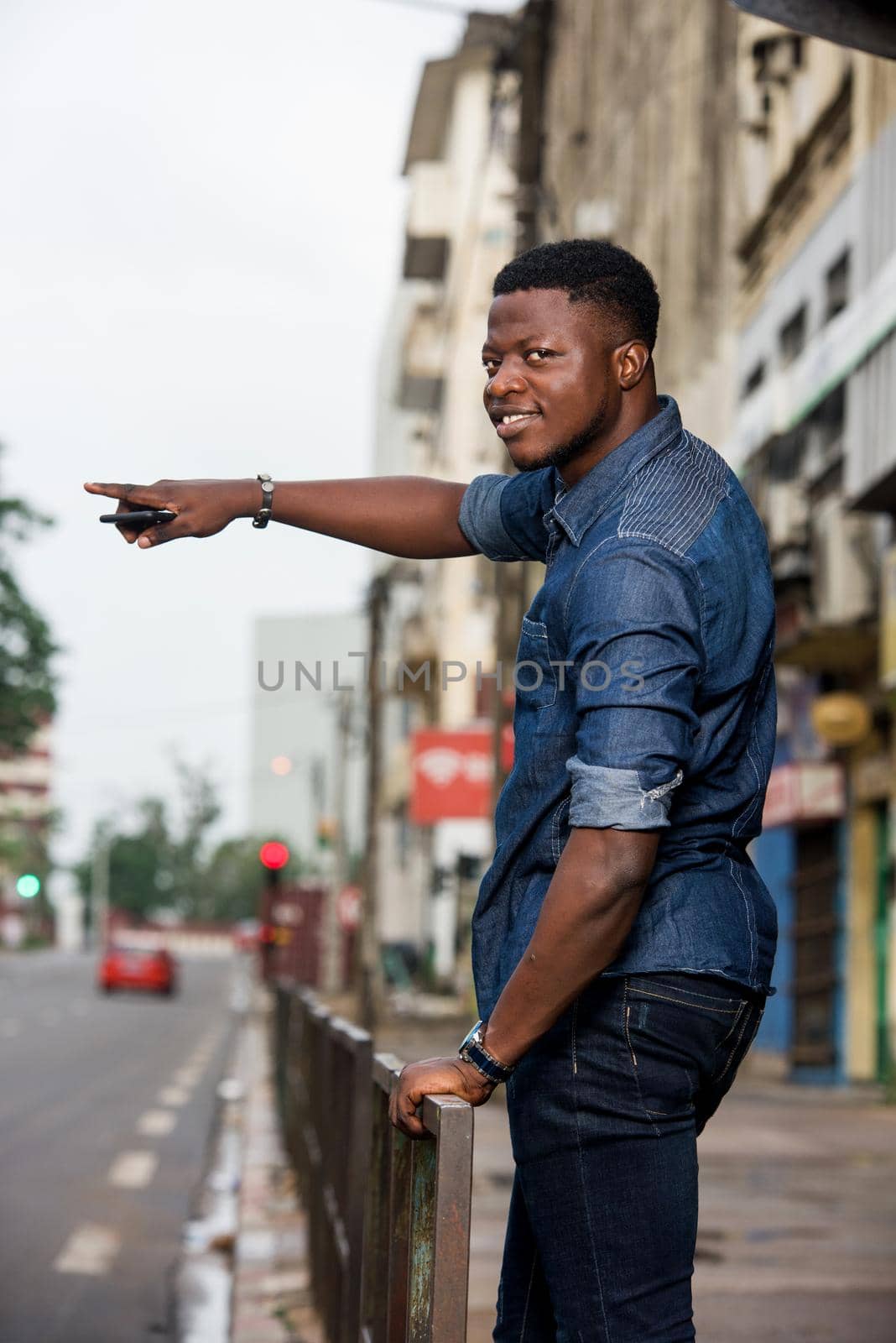 Young man smiling with his hand raised, holding a mobile phone and standing in the street waiting for taxi.