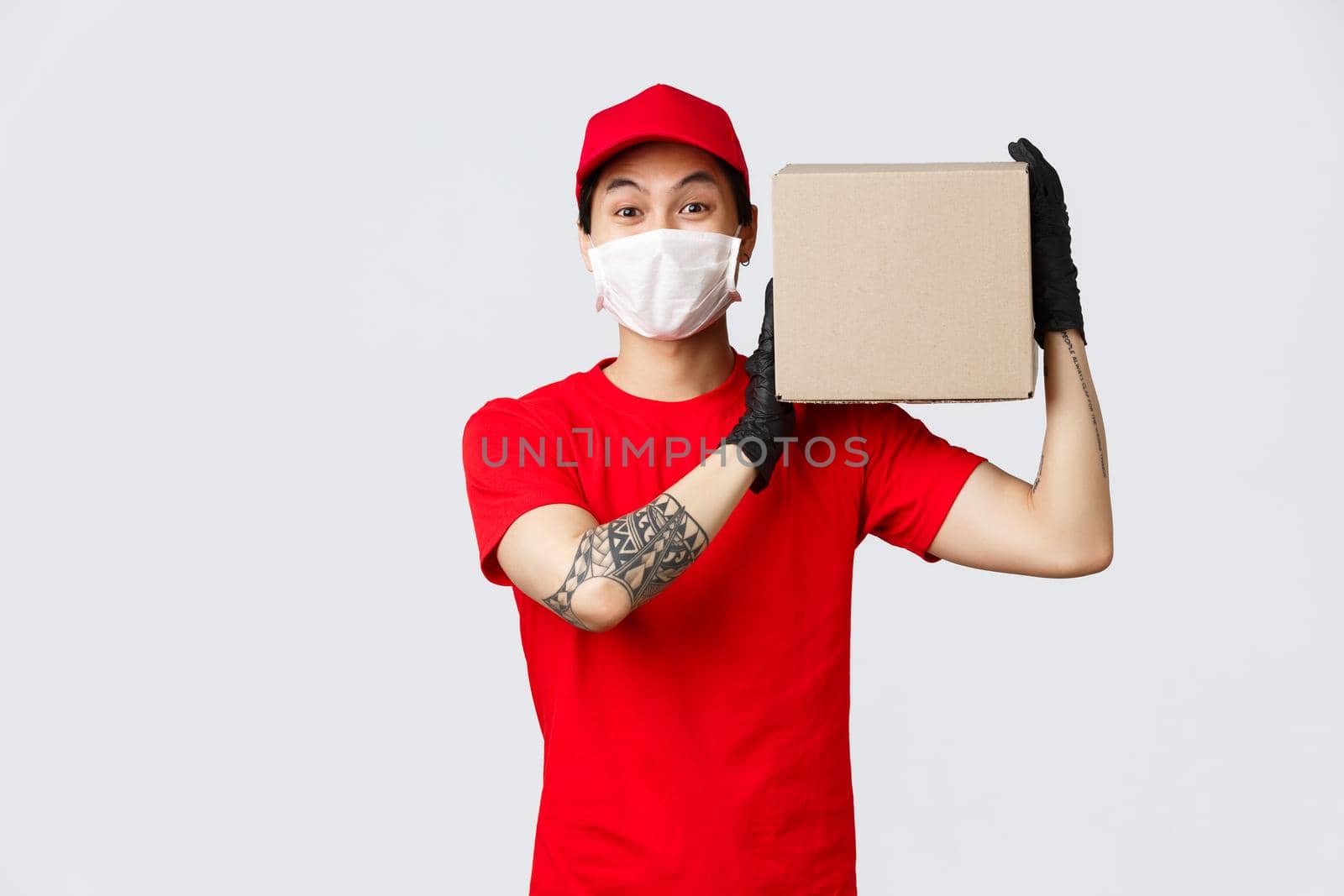 Concept of delivery during coronavirus pandemic. Charismatic asian courier holding package on shoulder, wear protective gloves and medical mask for client safety, give client their order.