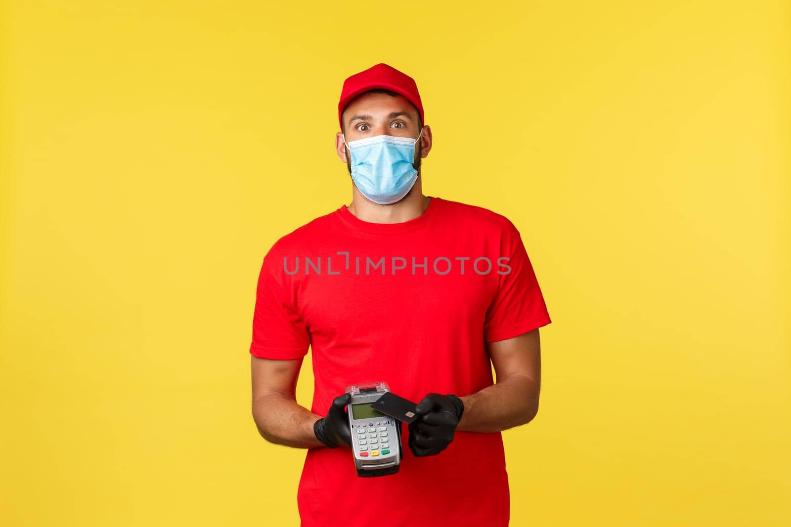 Food delivery, tracking orders, covid-19 and self-quarantine concept. Excited and amused courier in red uniform, medical mask and gloves, stare camera, using POS terminal and credit card.