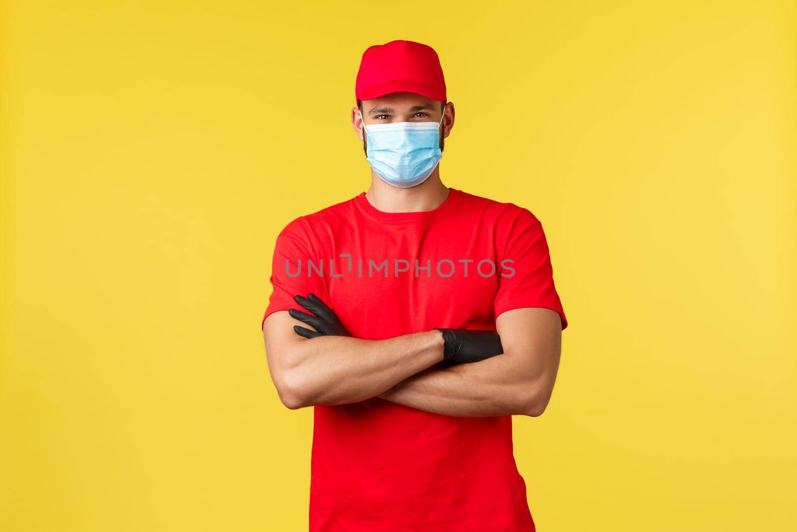 Express delivery during pandemic, covid-19, safe shipping, online shopping concept. Confident serious courier in red uniform, cross arms determined, wear medical mask, deliver orders to clients.