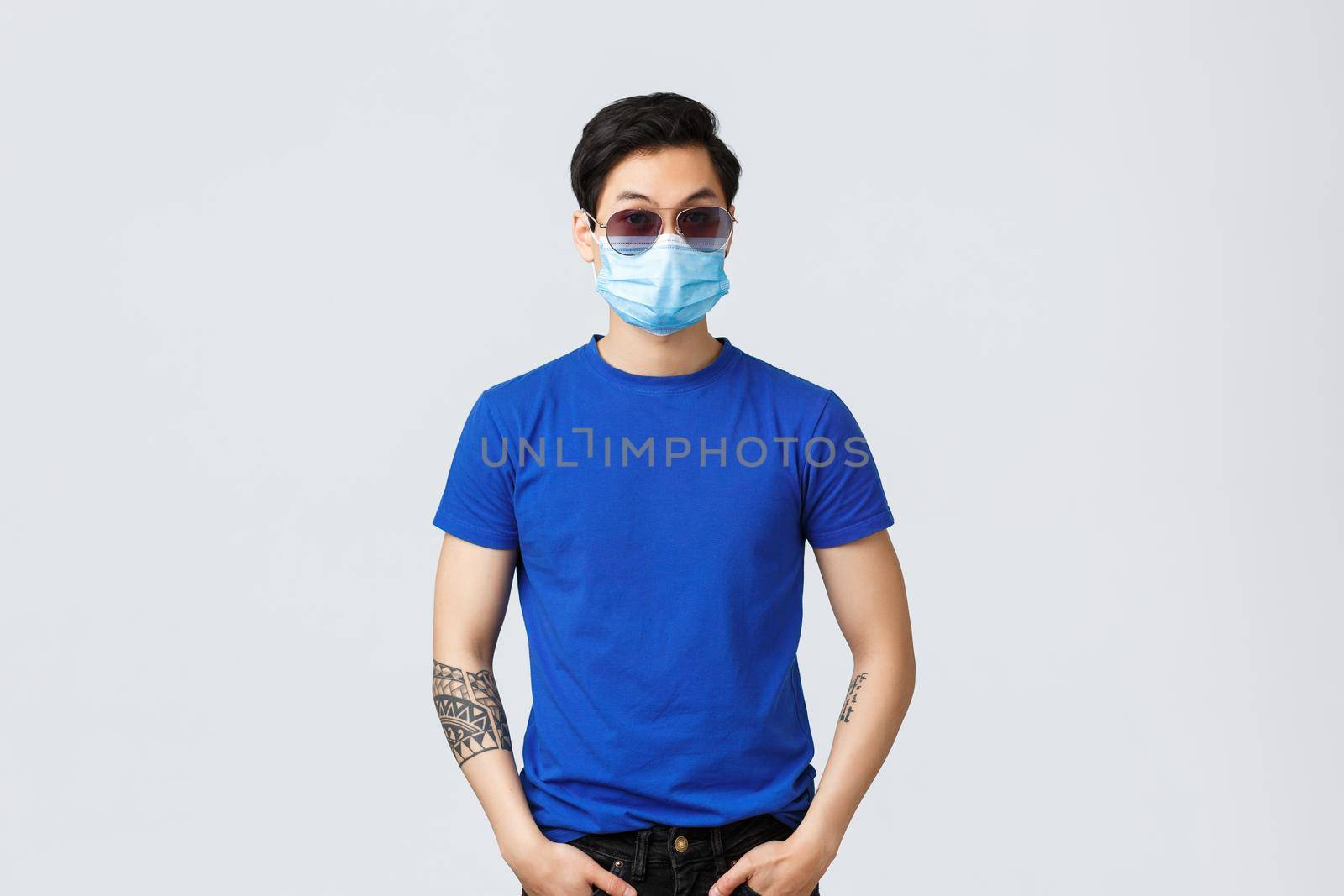 Covid019 lifestyle, people emotions and leisure on quarantine concept. Sassy and cool asian guy in sunglasses and medical mask acting like security guard or macho man, stand grey background by Benzoix