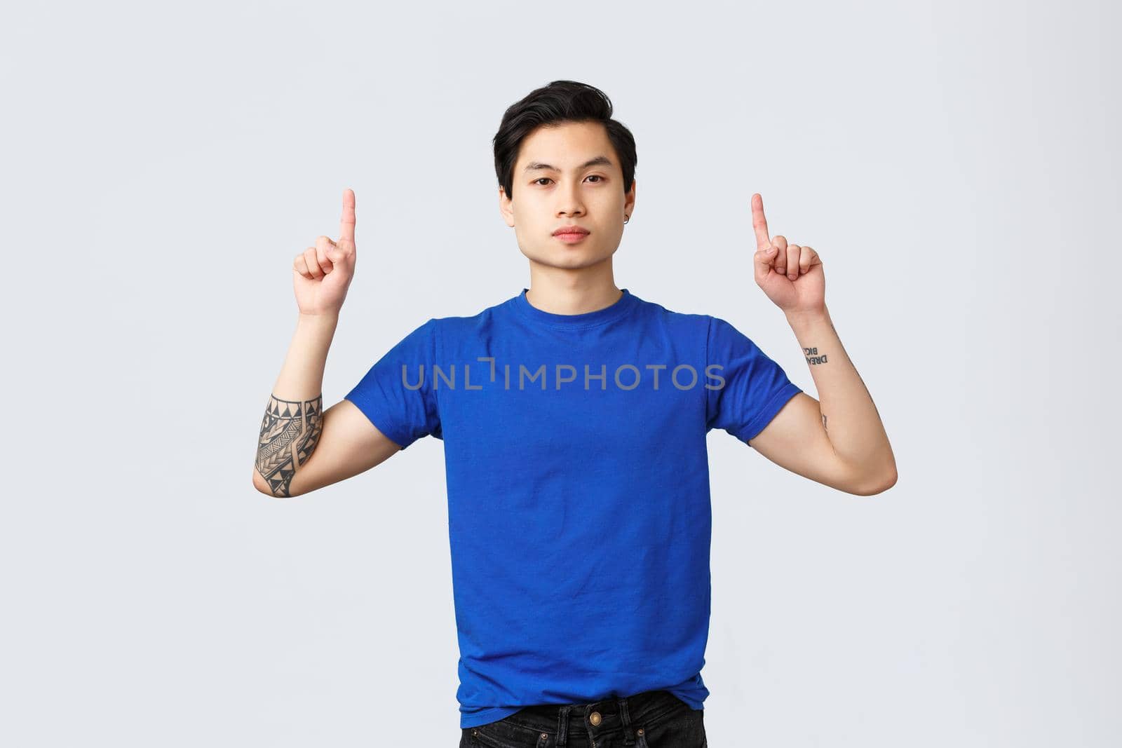 Different emotions, people lifestyle and advertising concept. Confident, serious asian man in blue t-shirt with tattoos, pointing fingers up, inform clients, showing way to banner, grey background.