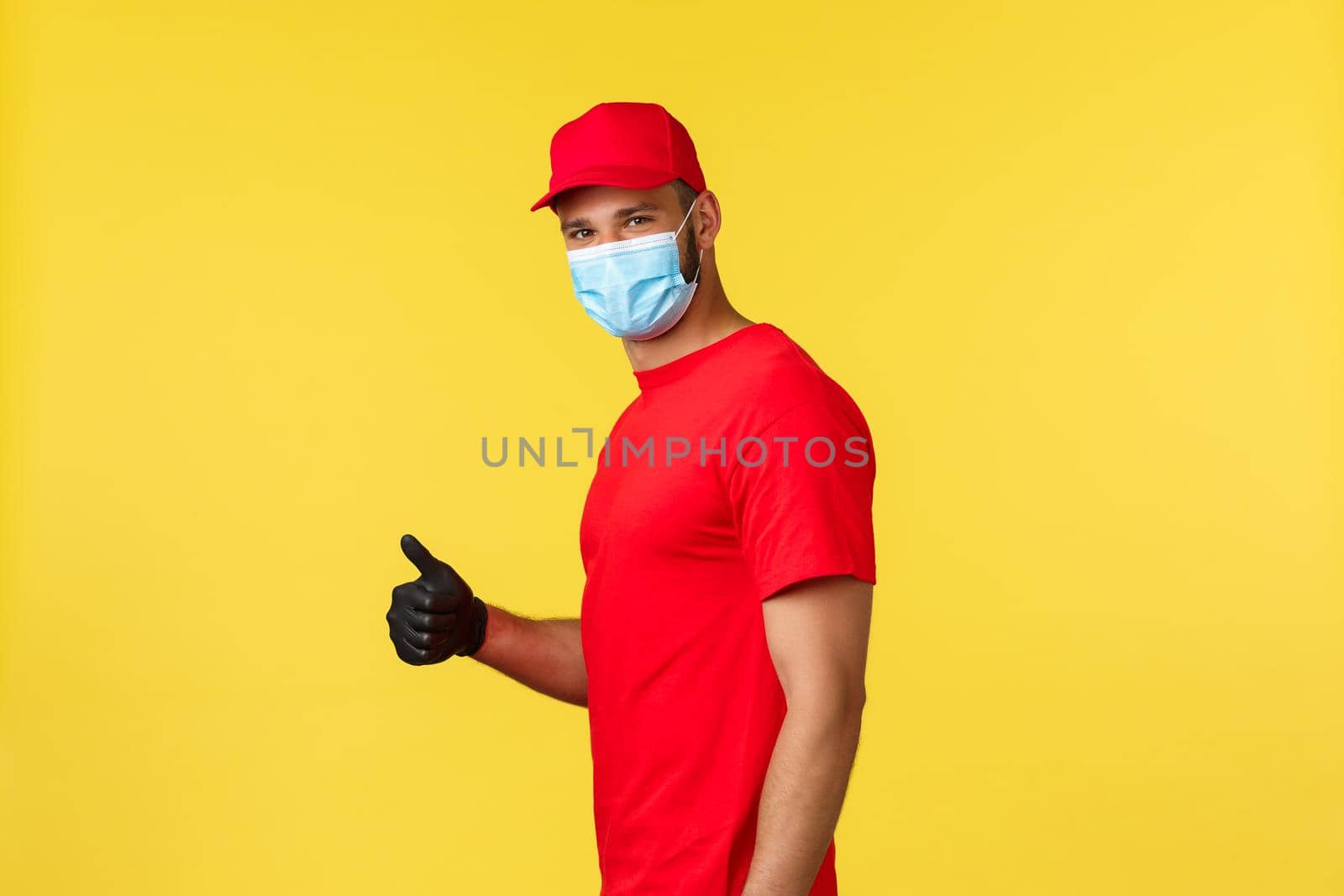 Express delivery during pandemic, covid-19, safe shipping, online shopping concept. Smiling handsome courier in red uniform and medical mask, show thumb-up, recommend package deliver service.