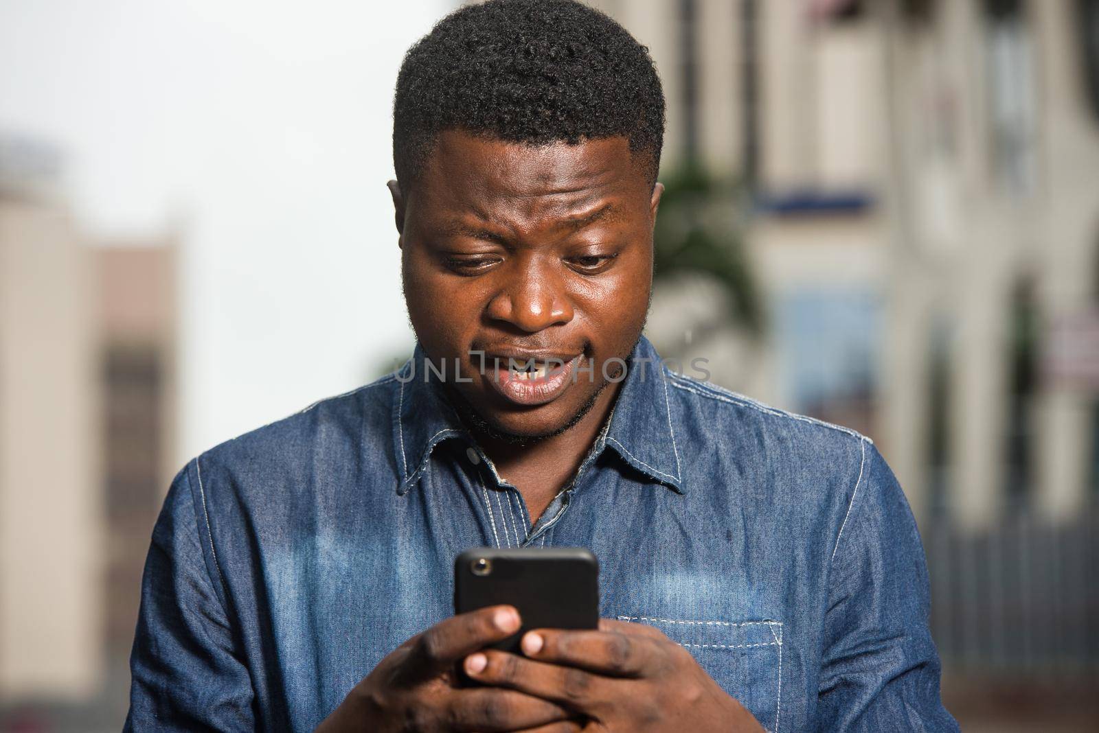 young african man standing in jeans shirt looking at mobile phone smiling.