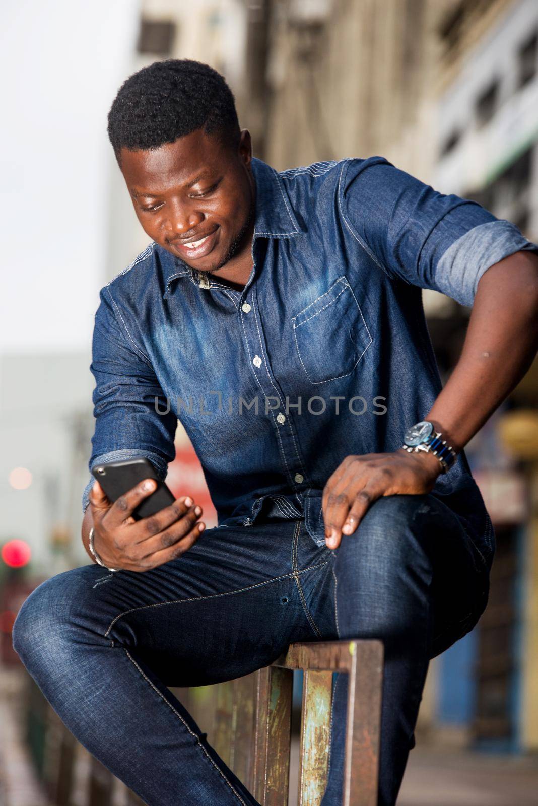 Portrait of a smiling young man sitting with a mobile phone on an iron bar in the street